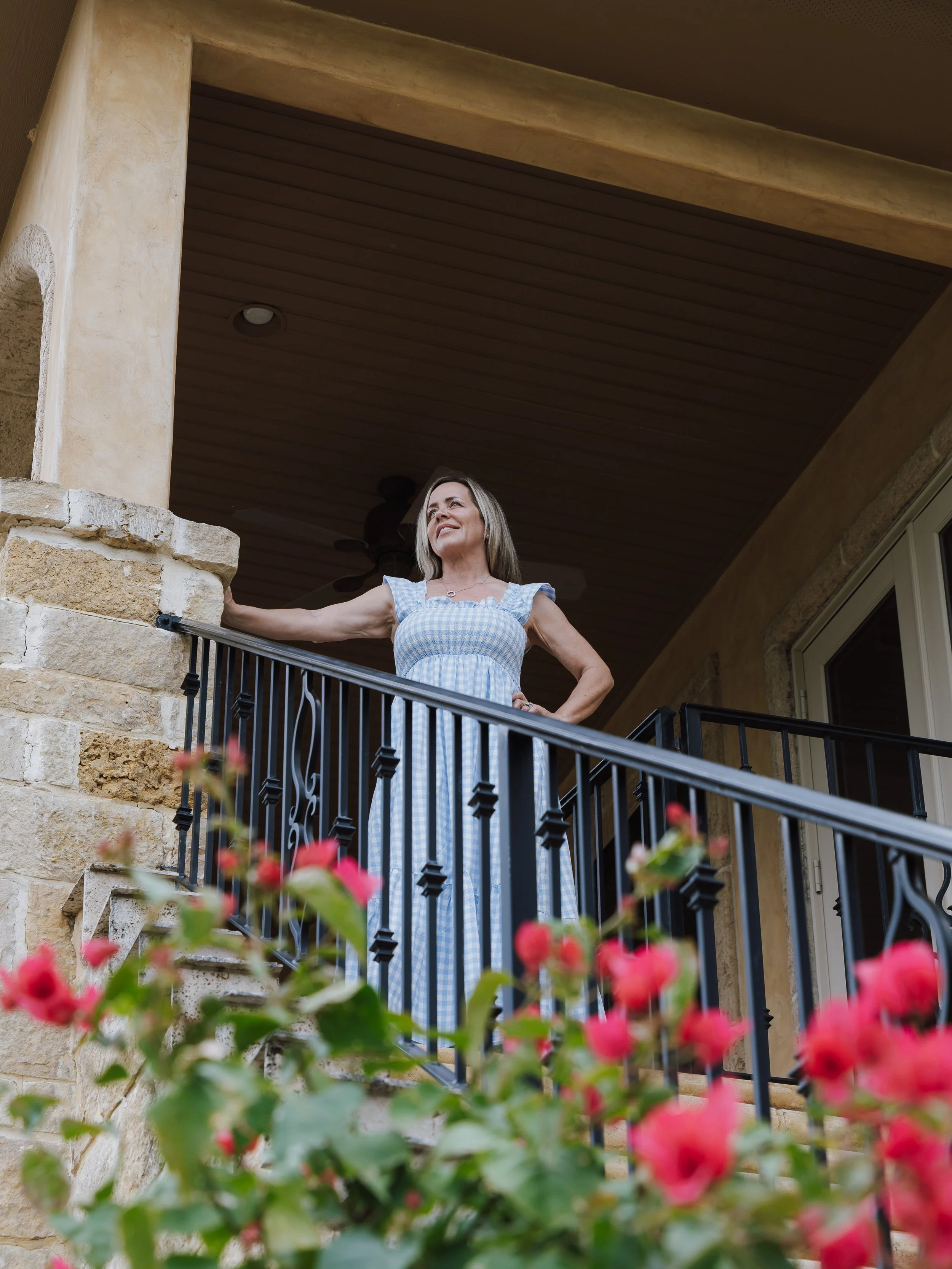 A middle-aged woman standing on a balcony, smiling and looking into the distance. There are flowers in the foreground and a stone and wood architectural background.