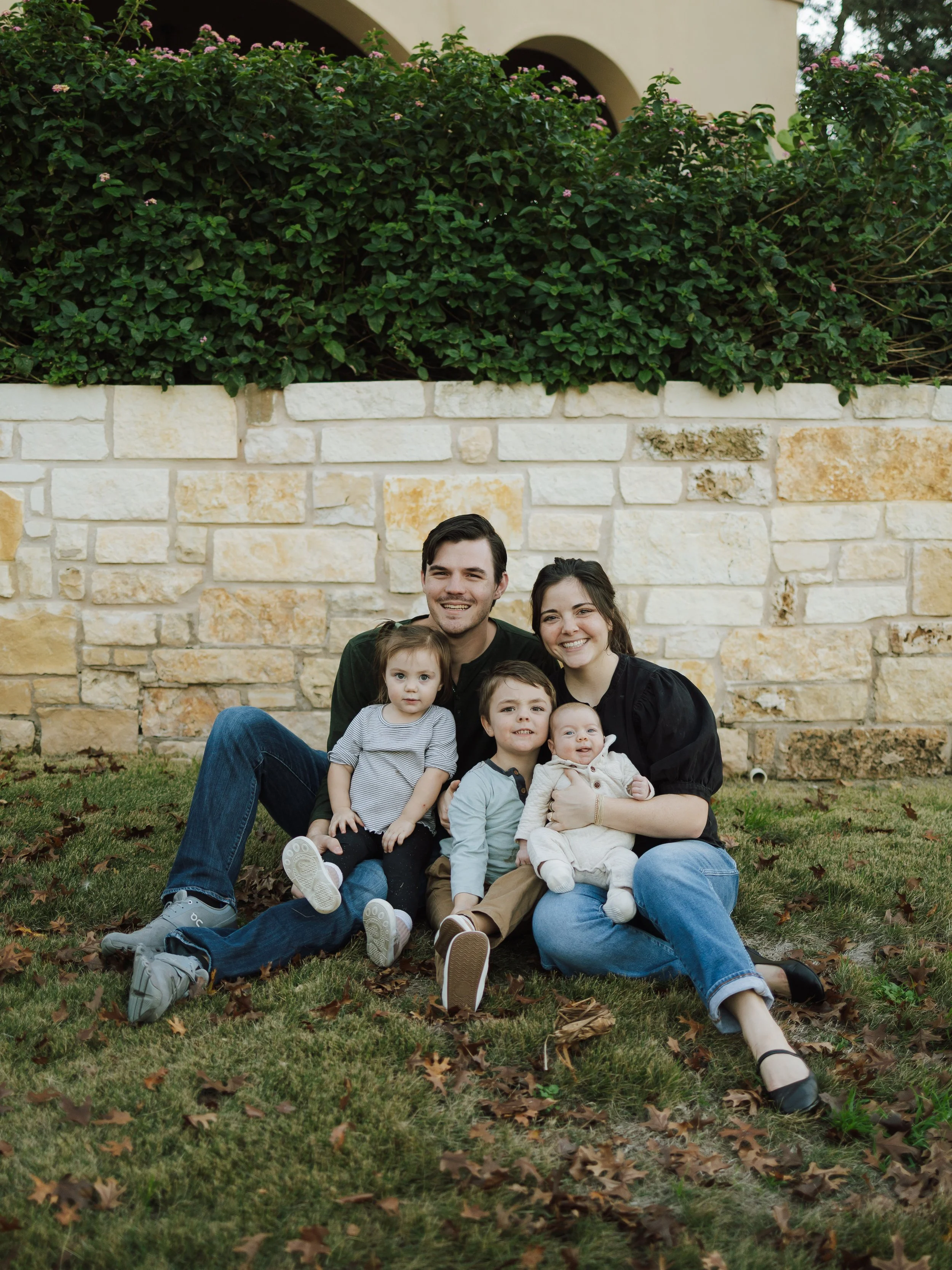 A family sitting on the grass in front of a stone wall, smiling at the camera. They include two adults and four children, with one young child sitting on the father's lap, and the other children around them. The background features a green hedge and part of a beige building with arches.