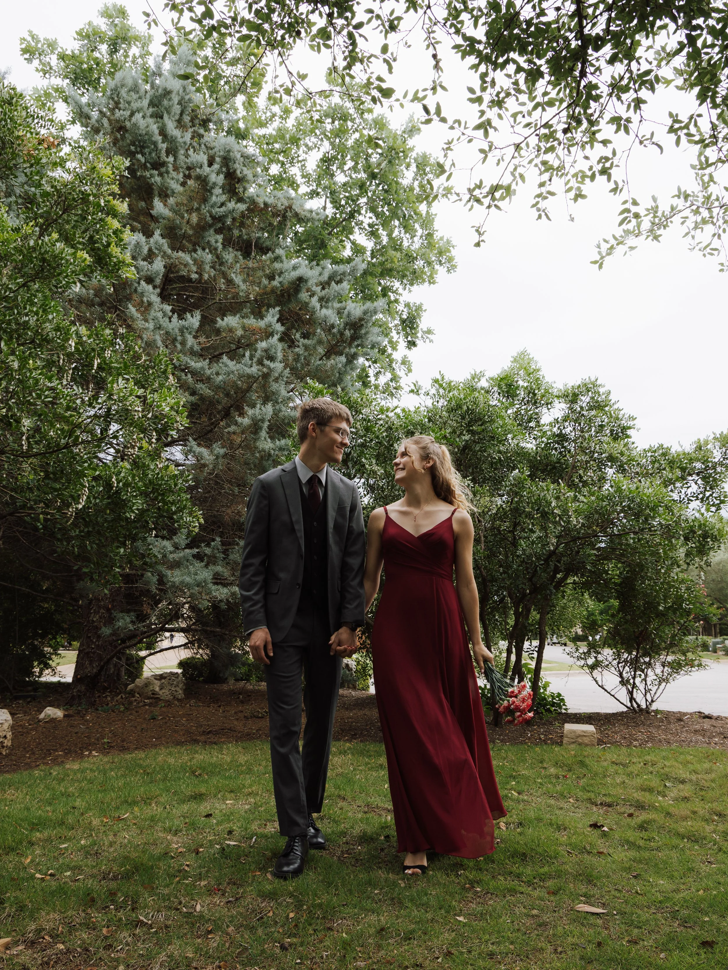 A couple dressed in formal attire walking hand in hand in a park with trees. The woman is holding a bouquet of flowers and wearing a long burgundy dress, while the man is in a dark suit and tie, looking at each other and smiling.