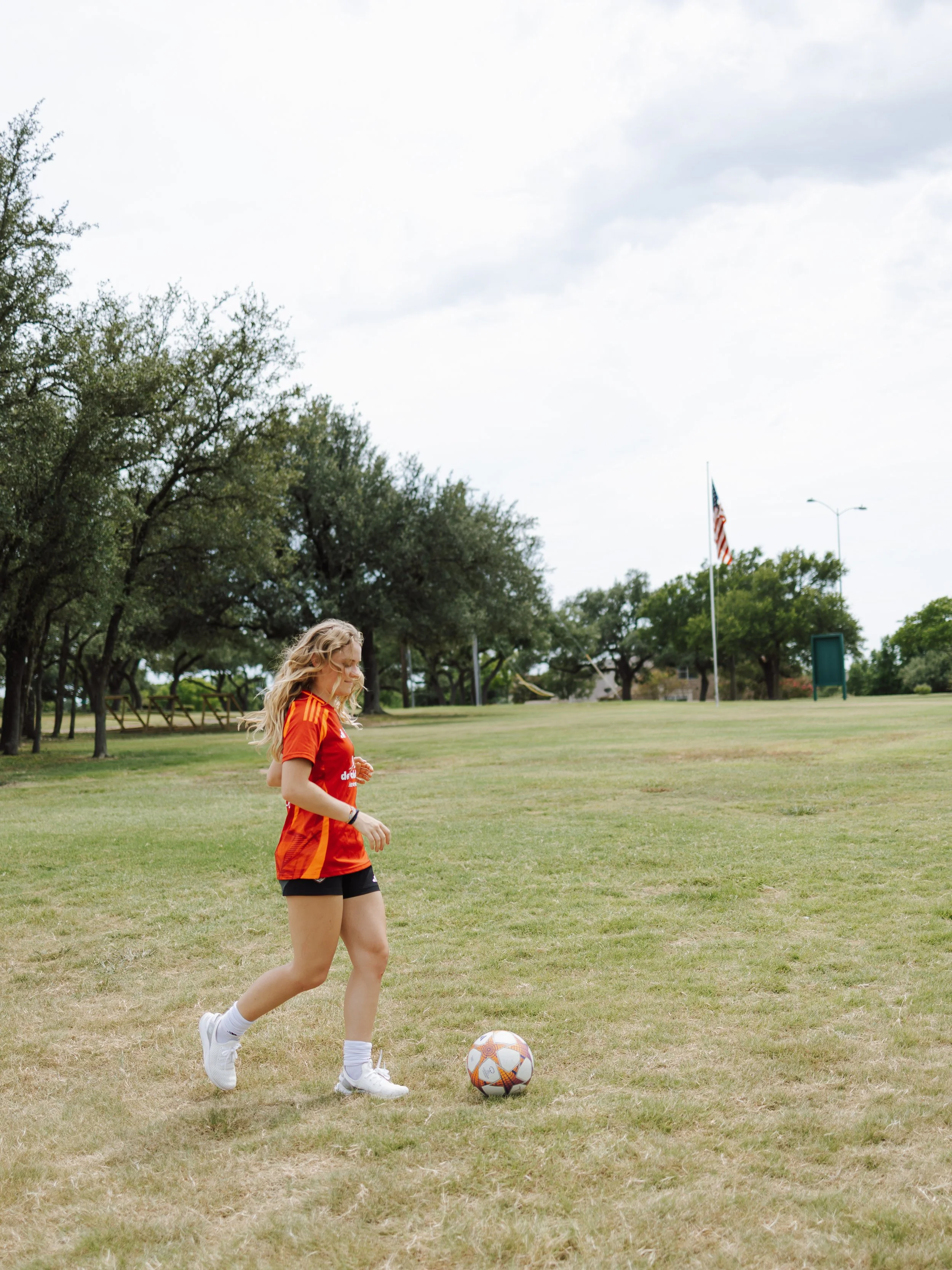 A young woman in a red soccer jersey and black shorts dribbles a soccer ball on a grassy field with trees and an American flag in the background.