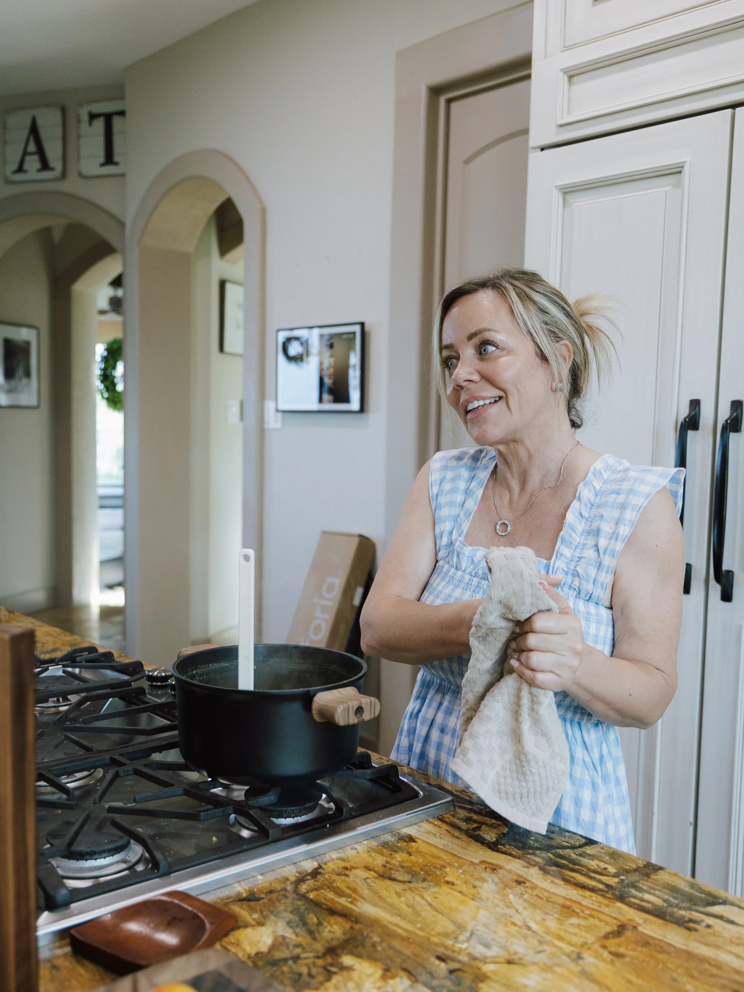 A woman in a blue checkered apron holding a kitchen towel, standing near a stove with a black pot, in a kitchen with beige walls and a granite countertop. There are decorative framed pictures on the wall and cabinets behind her.