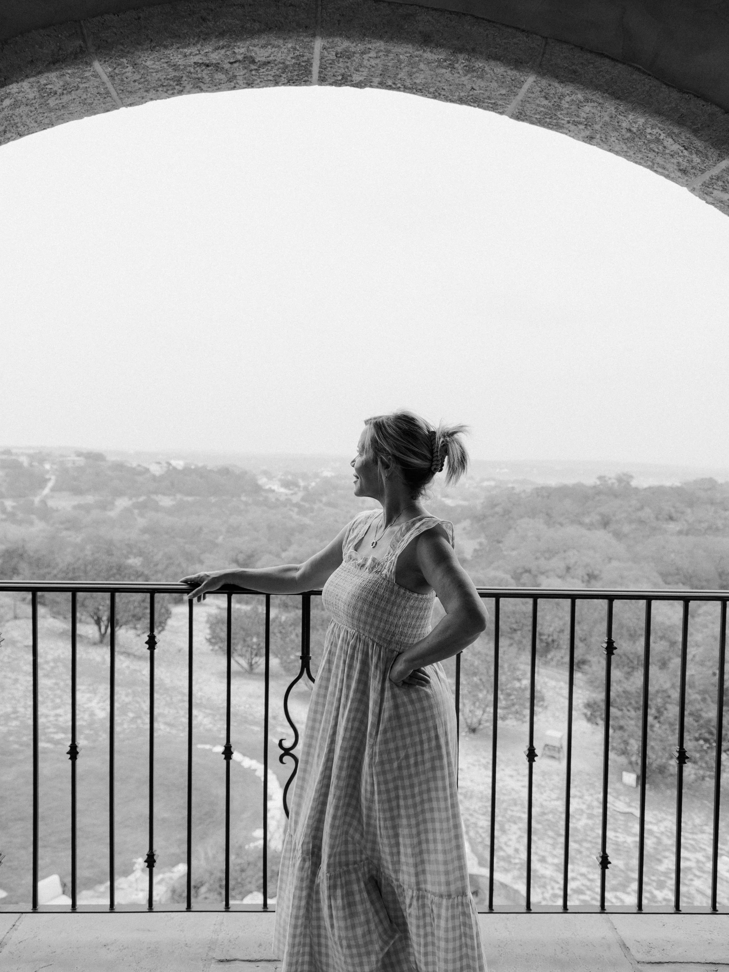 A woman stands on a balcony with a scenic landscape in the background, wearing a sleeveless dress and smiling.