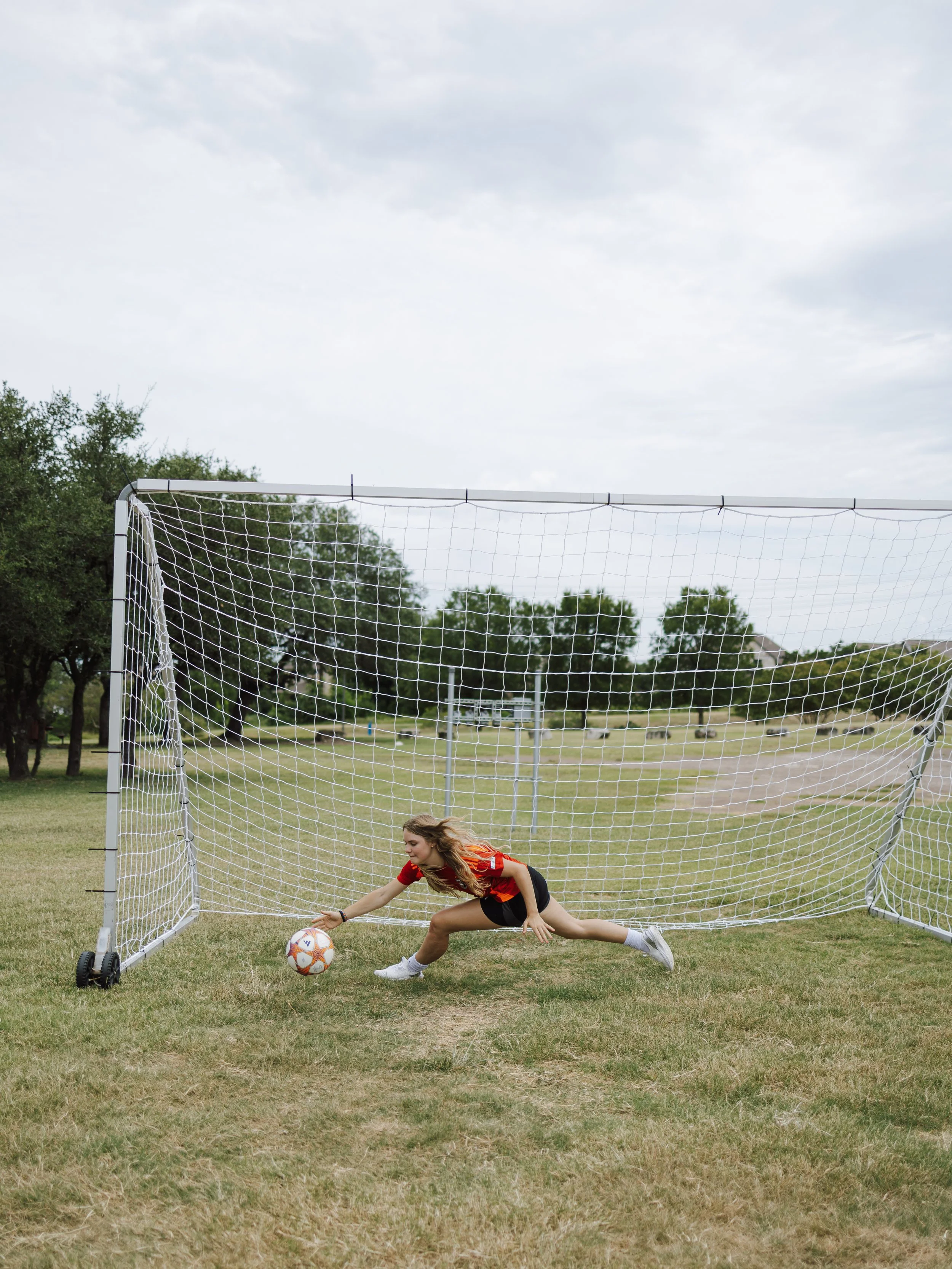 A young woman soccer player is diving to the ground to reach a soccer ball in front of a goal net, on a grassy field outdoors with trees and cloudy sky in the background.