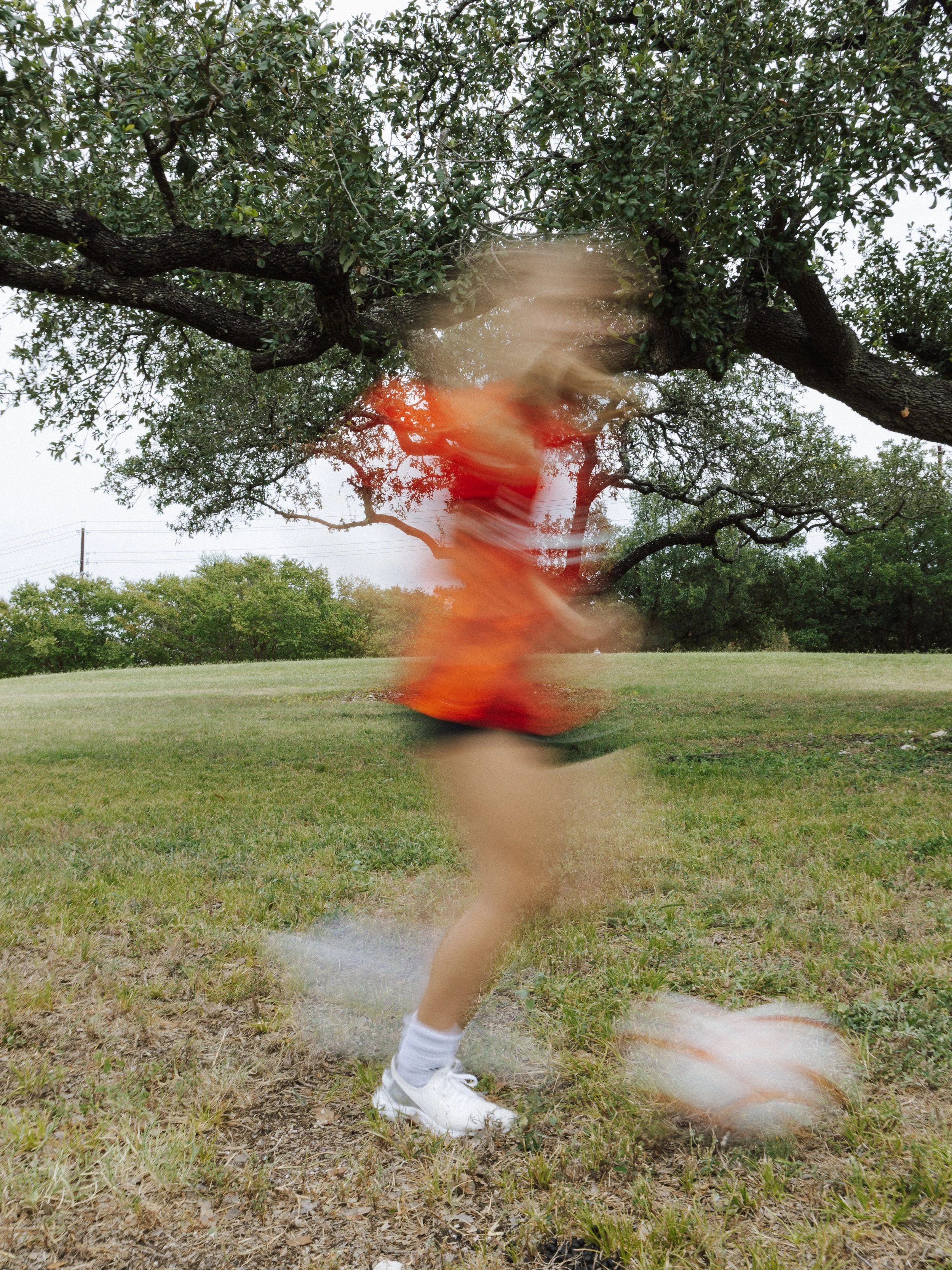 Blurred image of a person wearing white sneakers and orange clothing, jumping or falling on grass in a park-like setting with trees and cloudy sky in the background.