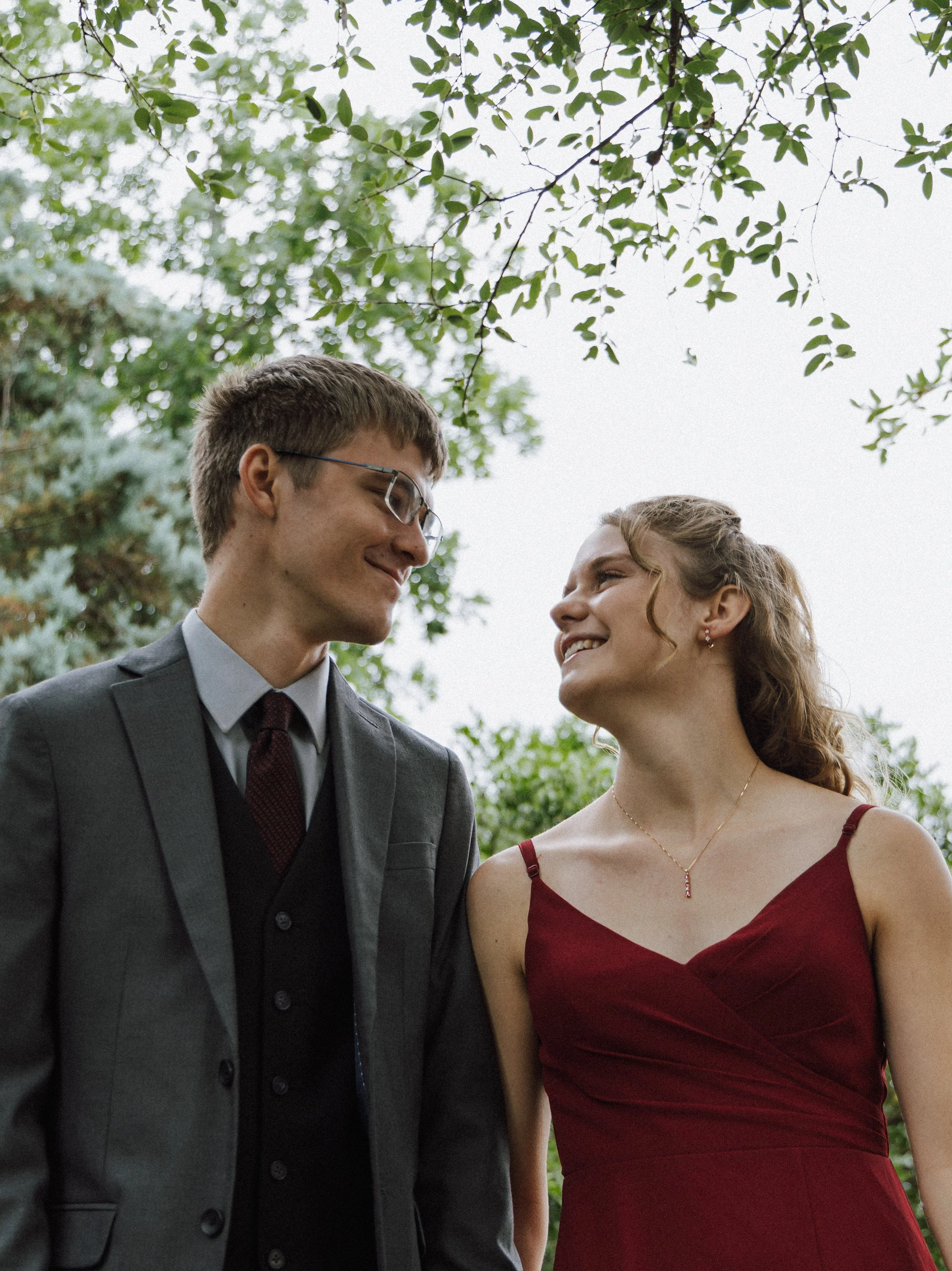 A young couple, dressed in formal attire, looking at each other outdoors beneath tree branches, smiling.