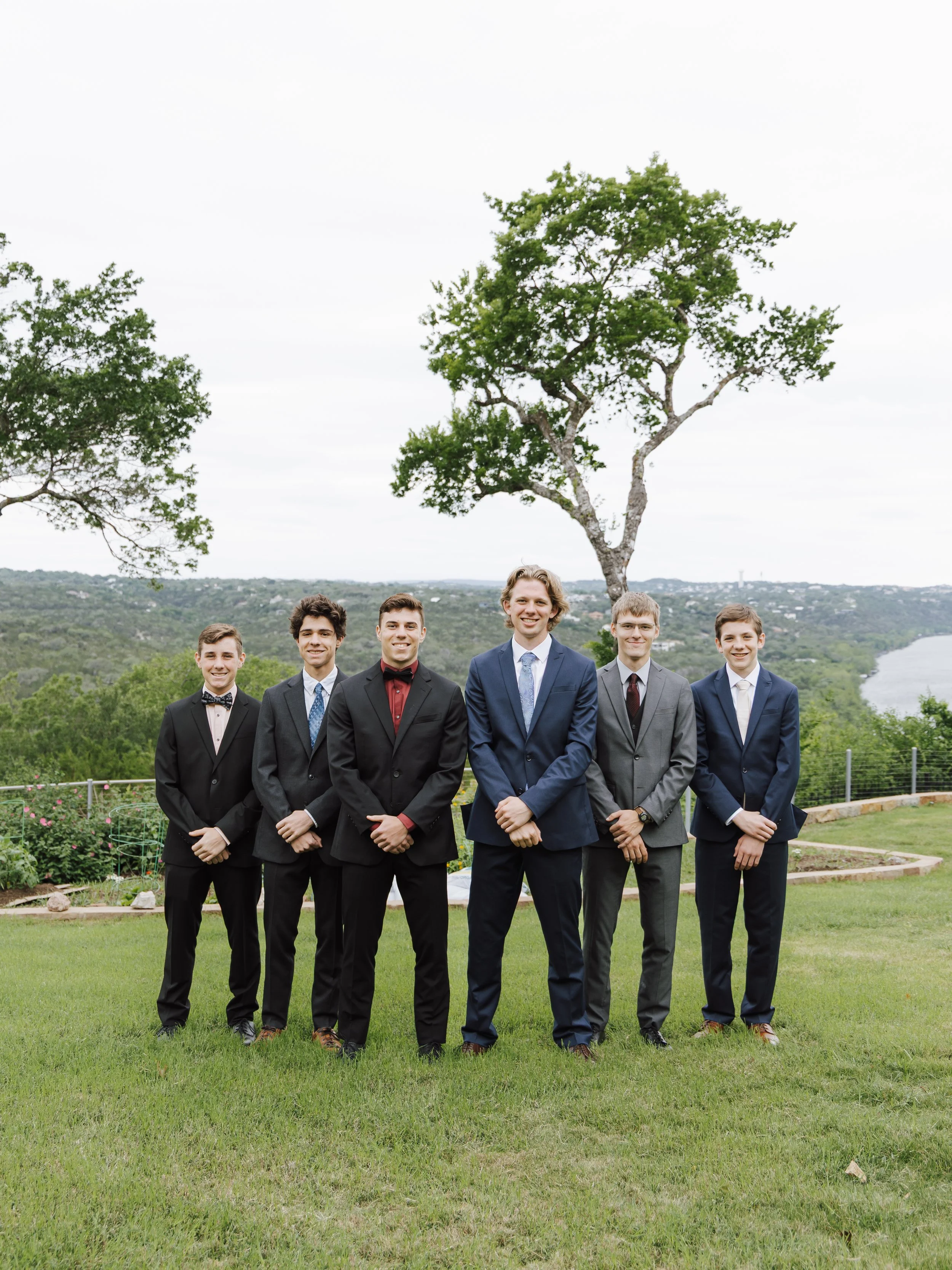 Six young men in suits standing outdoors on a lawn, smiling, with a large tree and a river in the background.