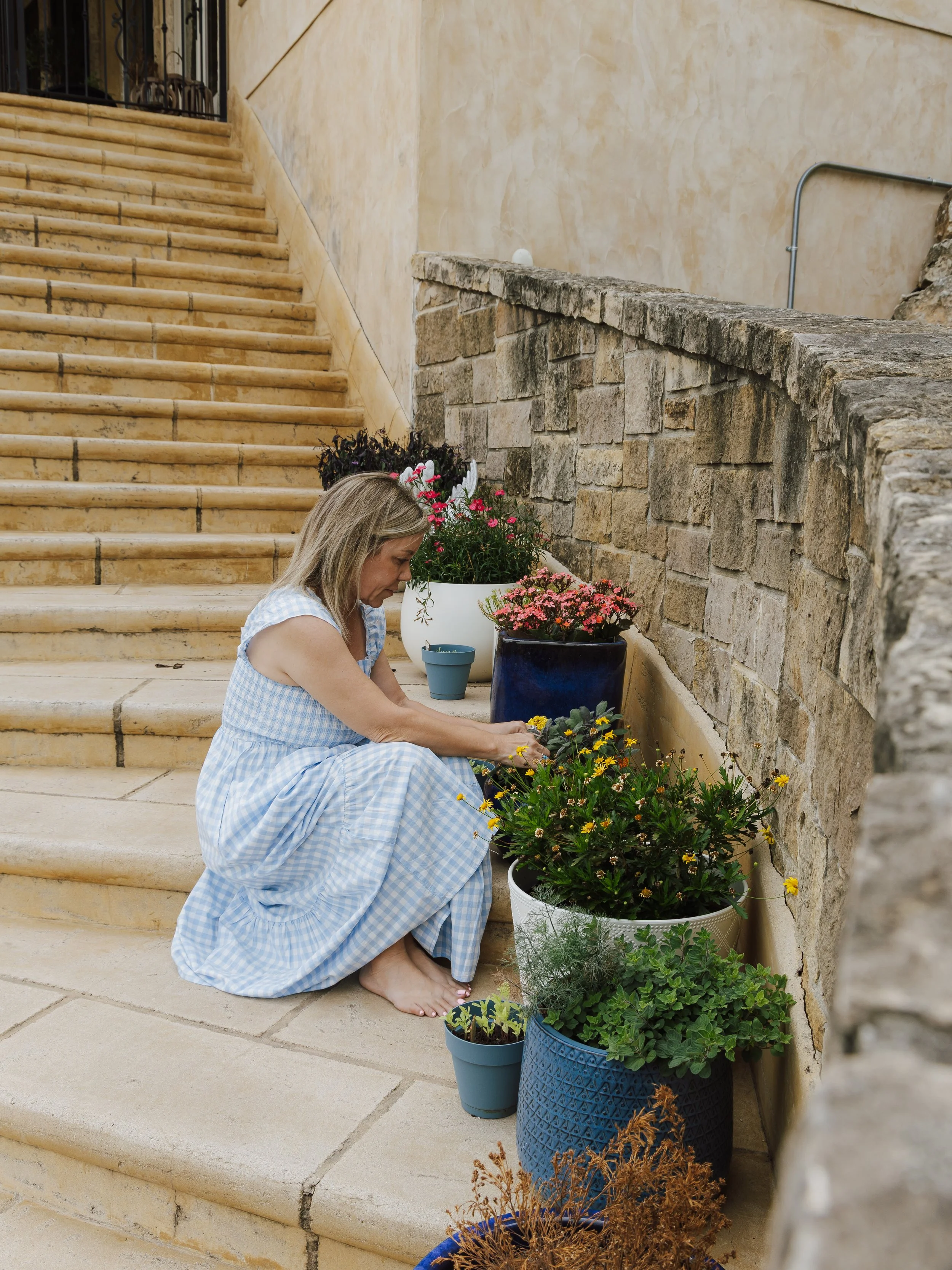 A woman is sitting barefoot on stone steps, tending to an array of colorful potted flowers against a stone and stucco wall.