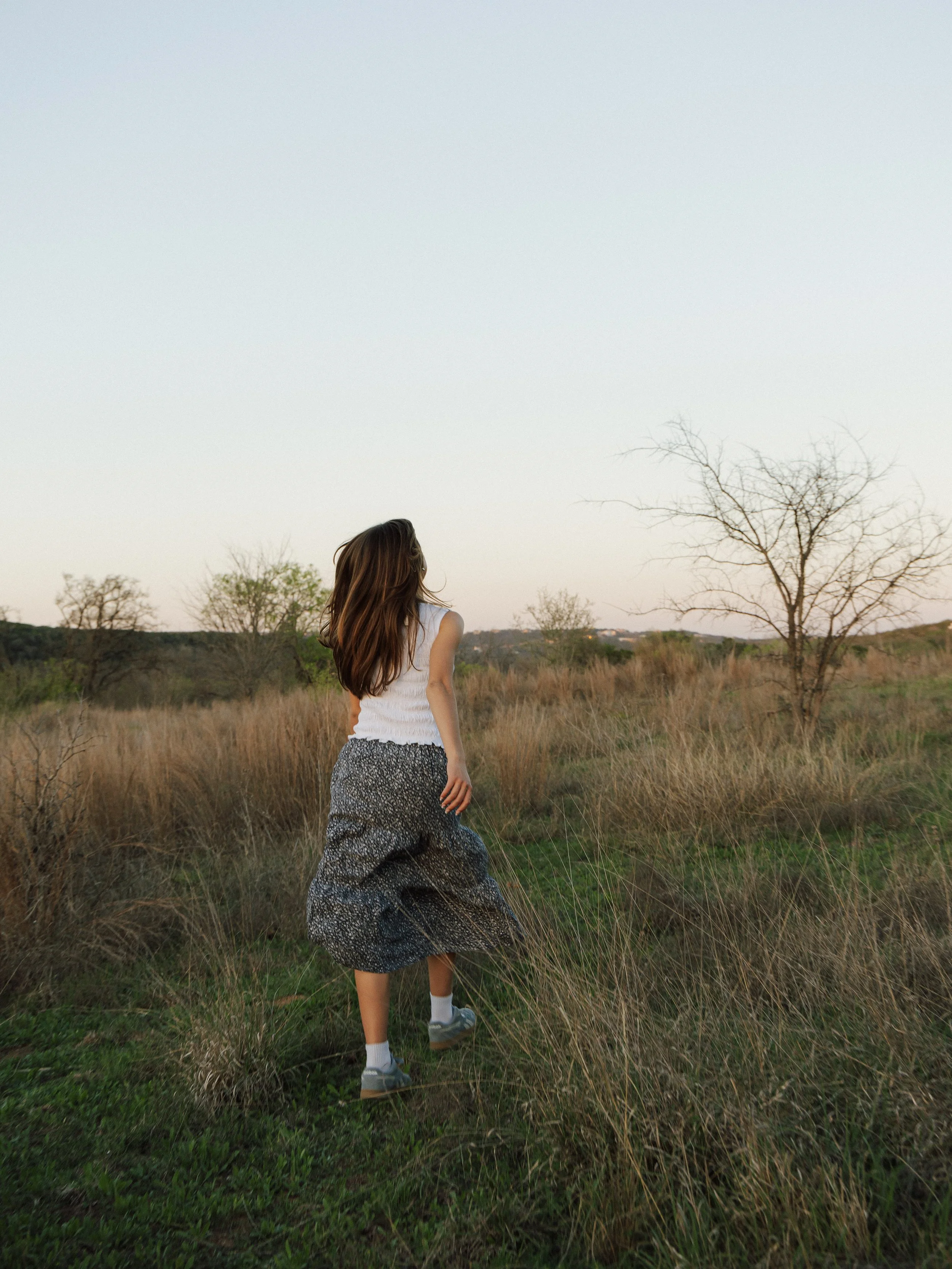 A girl walking through a grassy field with sparse trees, wearing a white sleeveless top and a long, patterned skirt during sunset.