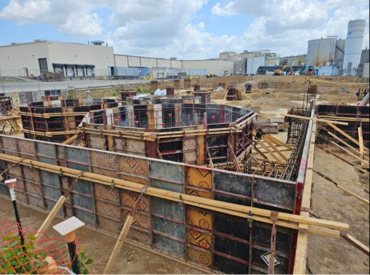 Construction site with walls being built for a structure, with formwork and rebar. Industrial buildings and storage tanks in the background under a partly cloudy sky.