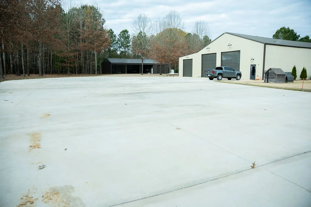 A large concrete driveway with a beige metal building that has three garage doors, a pickup truck parked outside, and a smaller dark-colored building with trees in the background.