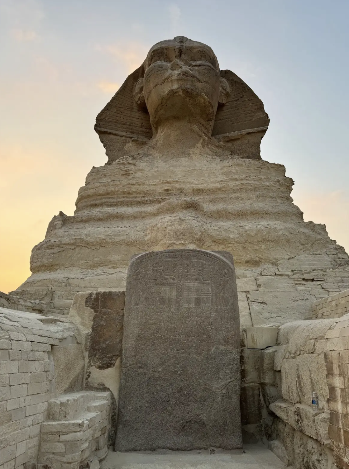 A large ancient stone statue of a pharaoh's bust at sunset, with an inscribed stone tablet in the foreground, surrounded by old brick ruins.