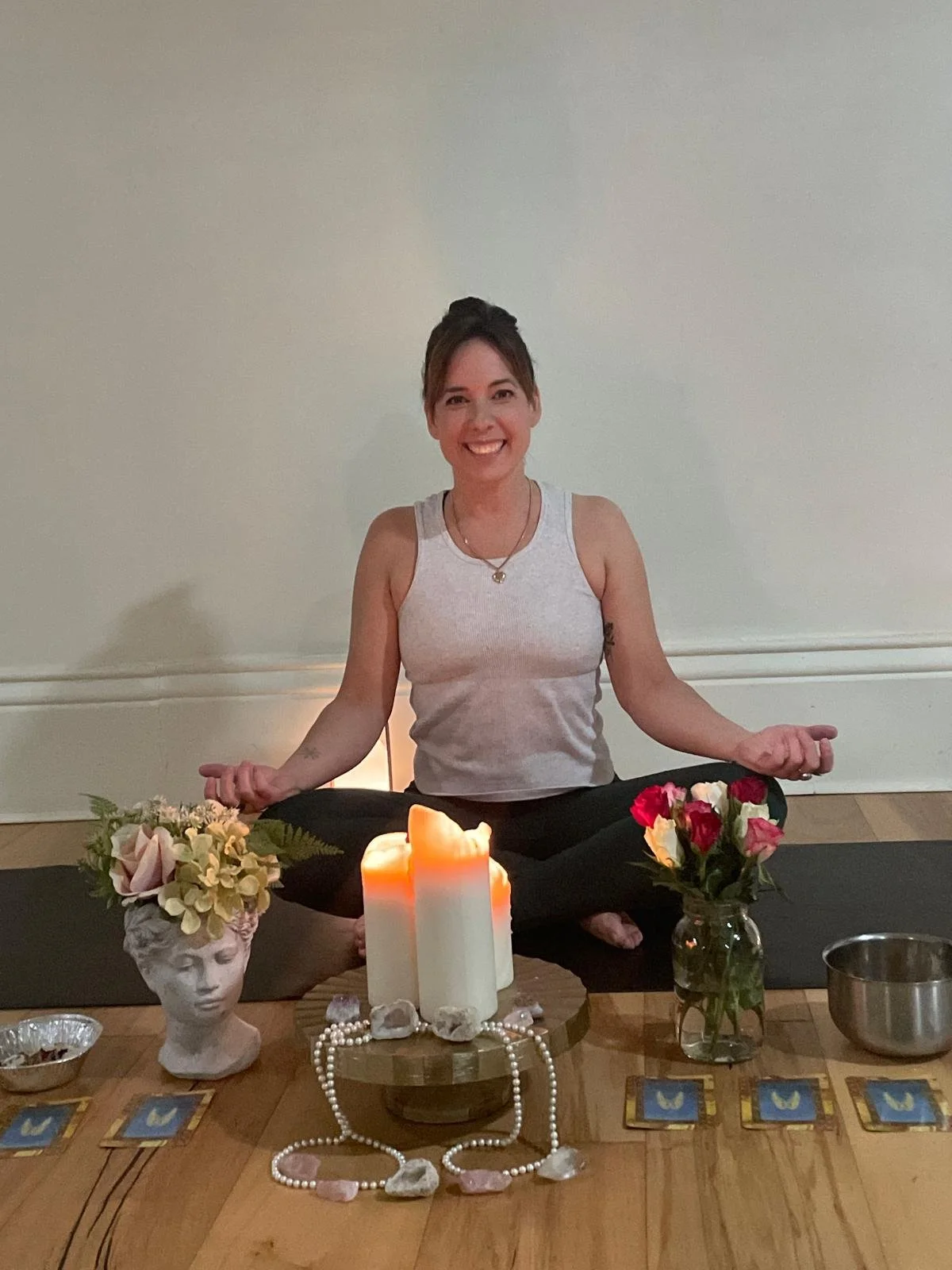 A woman sitting cross-legged on a yoga mat, smiling, surrounded by candles, flowers in vases, tarot cards, and crystals on a wooden table, engaging in a meditation or spiritual practice.