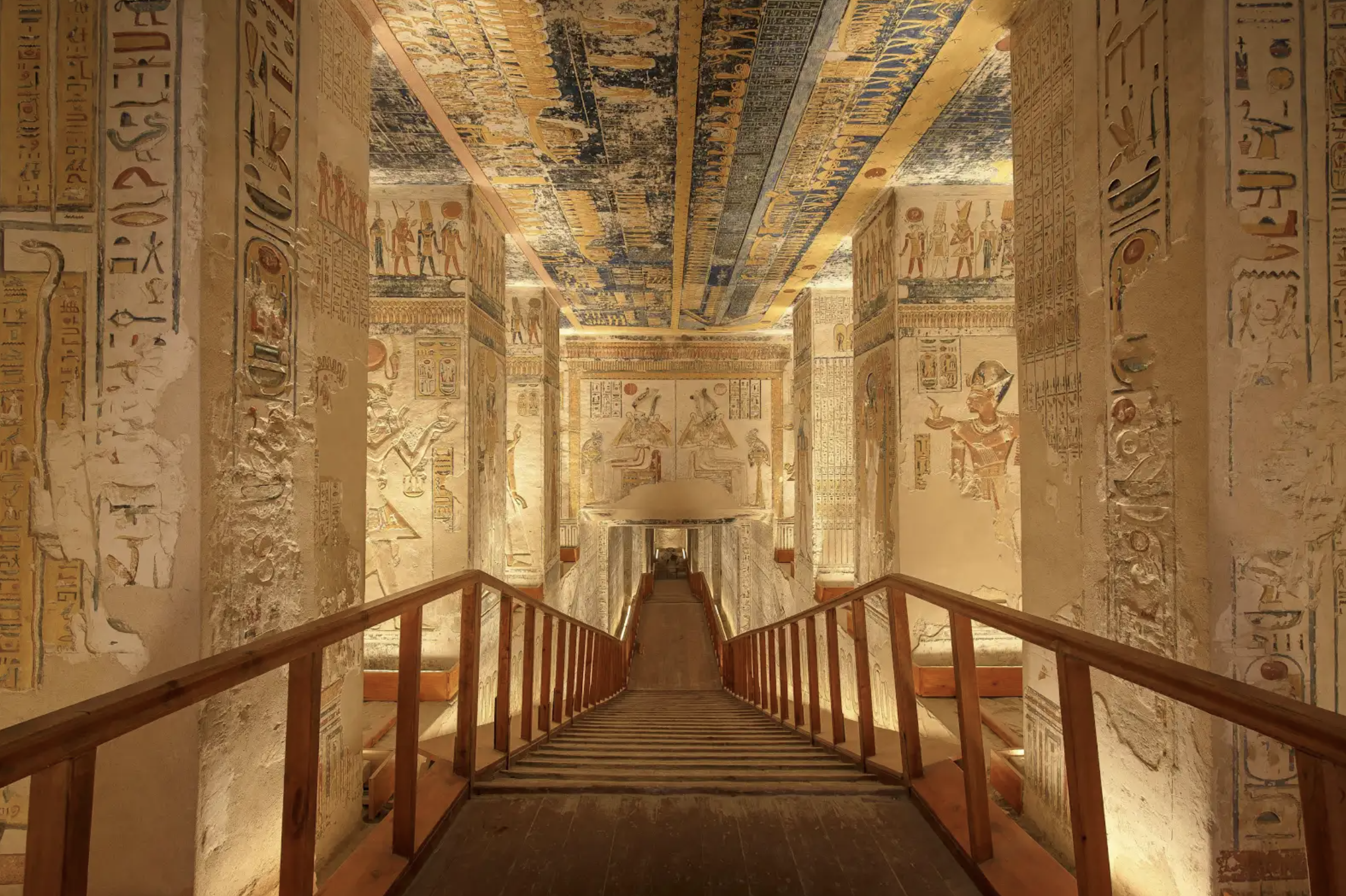 Interior view of ancient Egyptian tomb with hieroglyphic carvings and colorful murals on stone walls and ceiling, accessed by a wooden ramp.