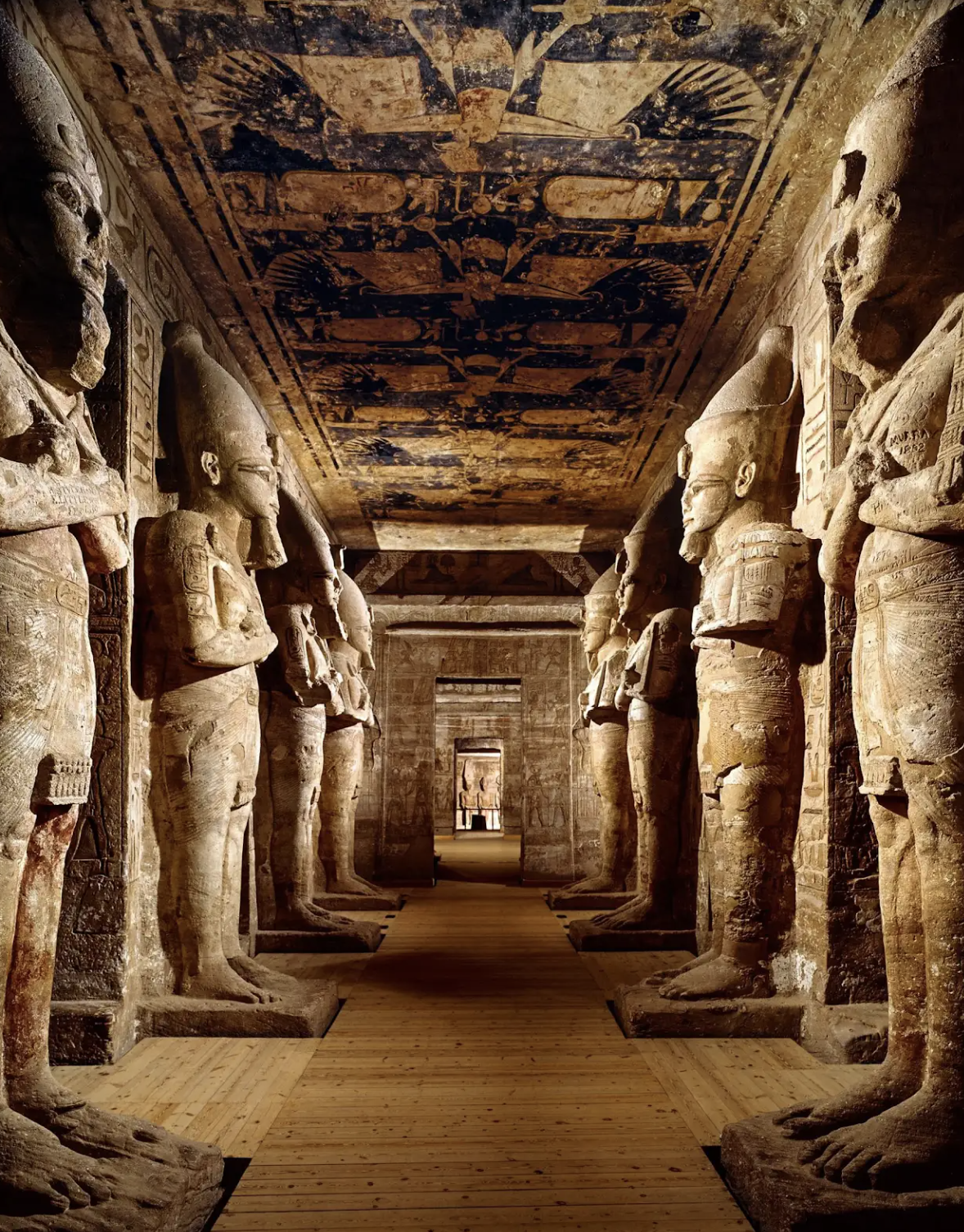 Ancient Egyptian statues and hieroglyphs inside a temple hallway with a wooden floor, featuring large statues lining both sides and a decorated ceiling.