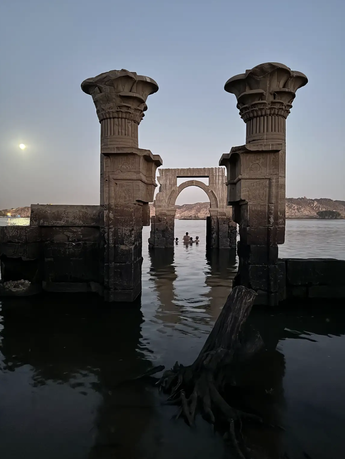 Ancient Egyptian-style stone structures with carved columns, partially submerged in water, with a few people swimming nearby during dusk or dawn.