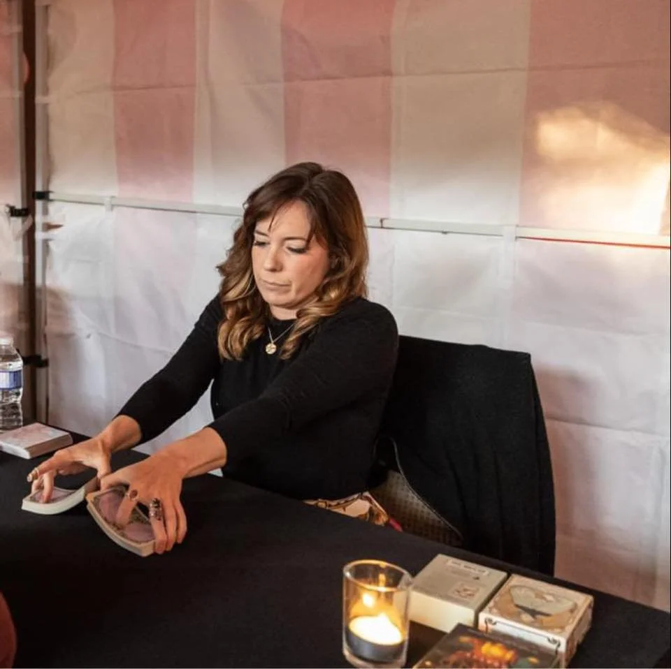 A woman sitting at a table, using tarot cards. The table has a water bottle, some books, a lit candle, and tarot card boxes. The background features a pink and white tent or partition.