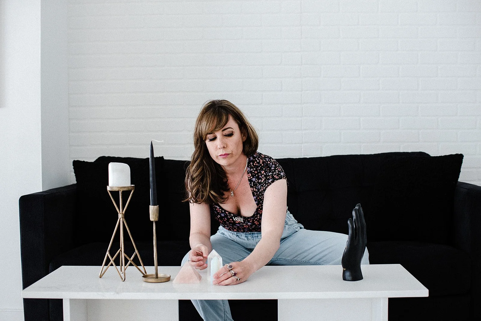 A woman with brown hair and a floral top sits on a black couch, arranging tarot cards on a white table, with decorative items including a candle, a black hand sculpture, and a black candle in a holder.