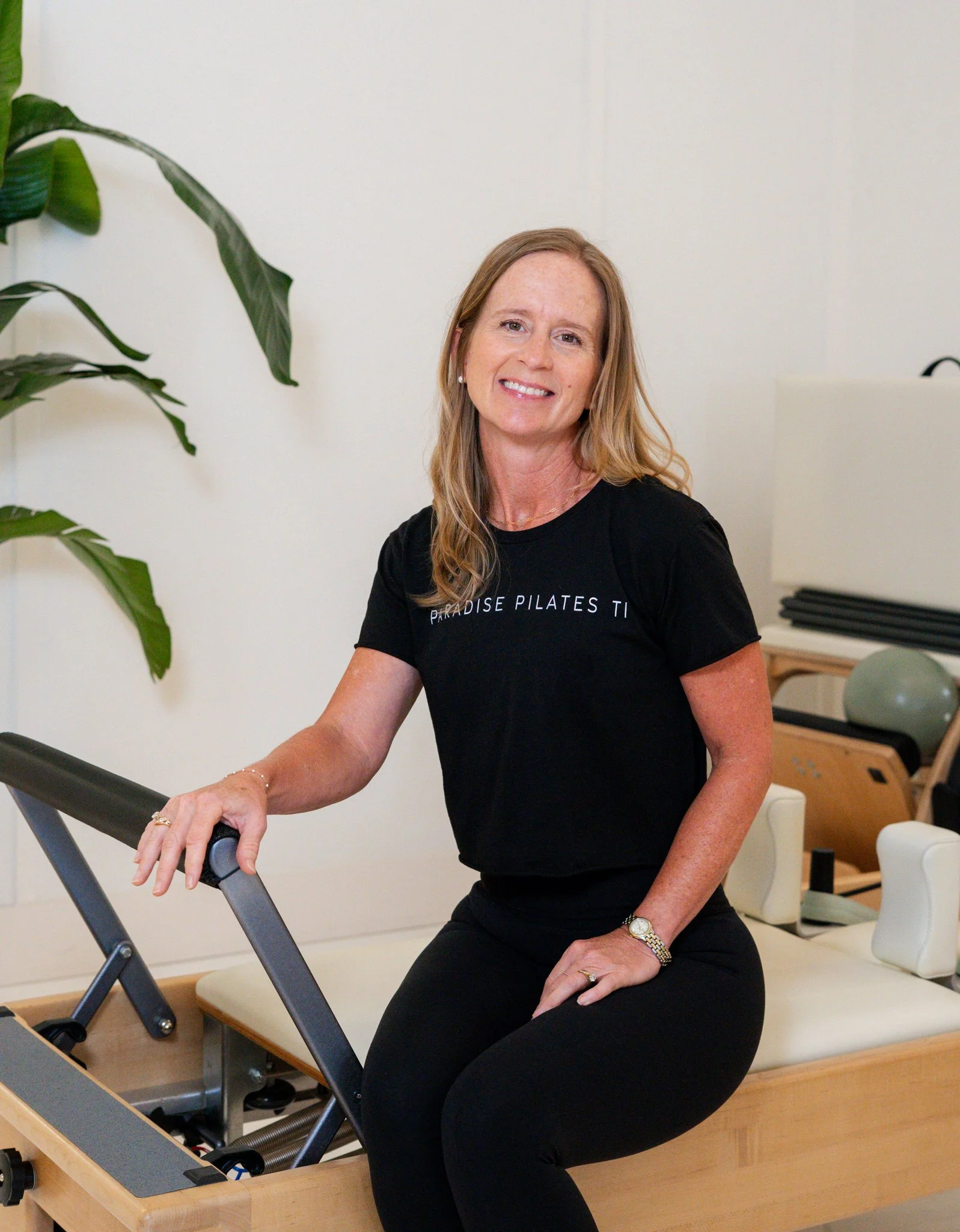 Smiling woman in a black shirt standing in a bright room with green plants.