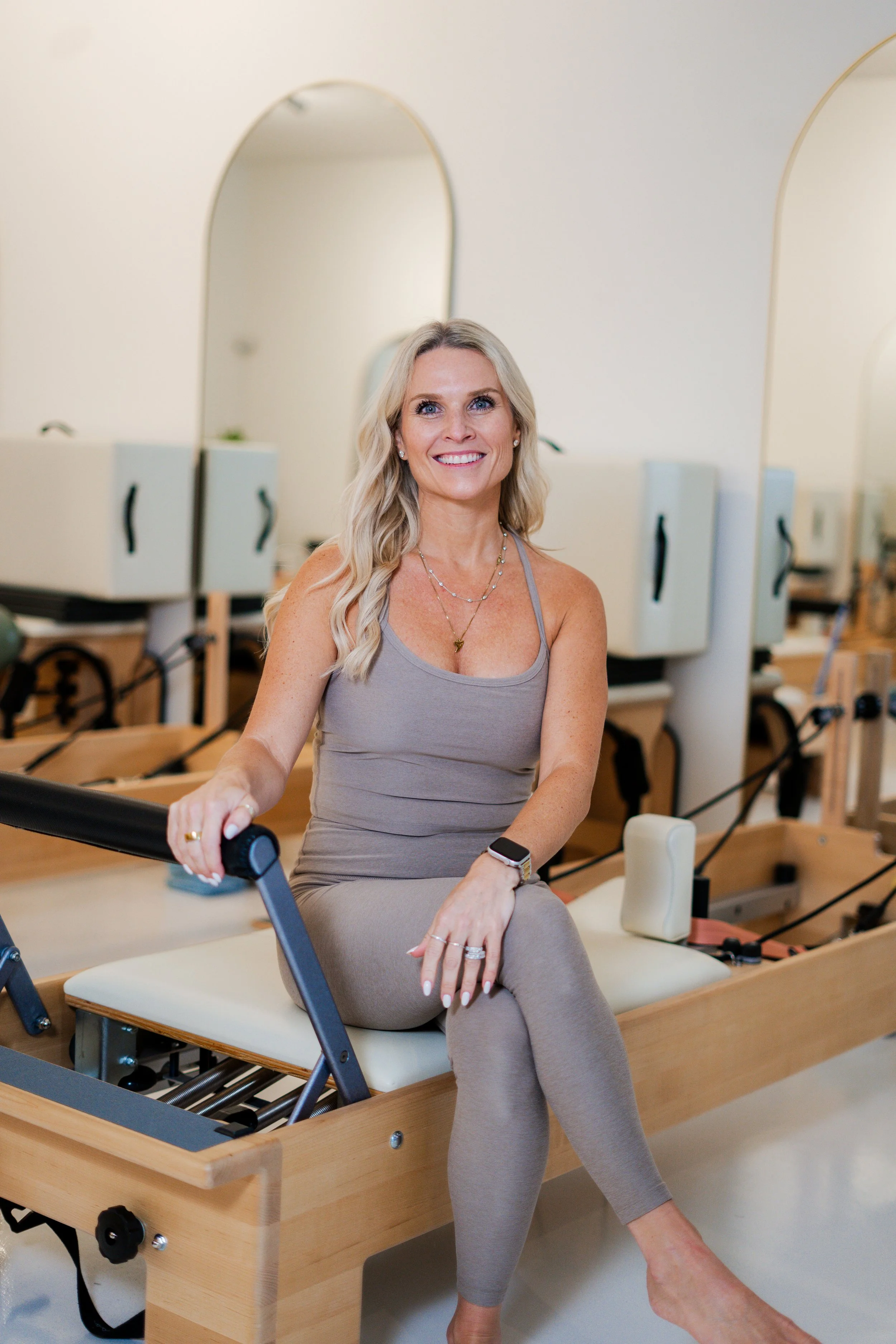 Woman sitting on the floor in a Pilates studio with reformer machines in the background.