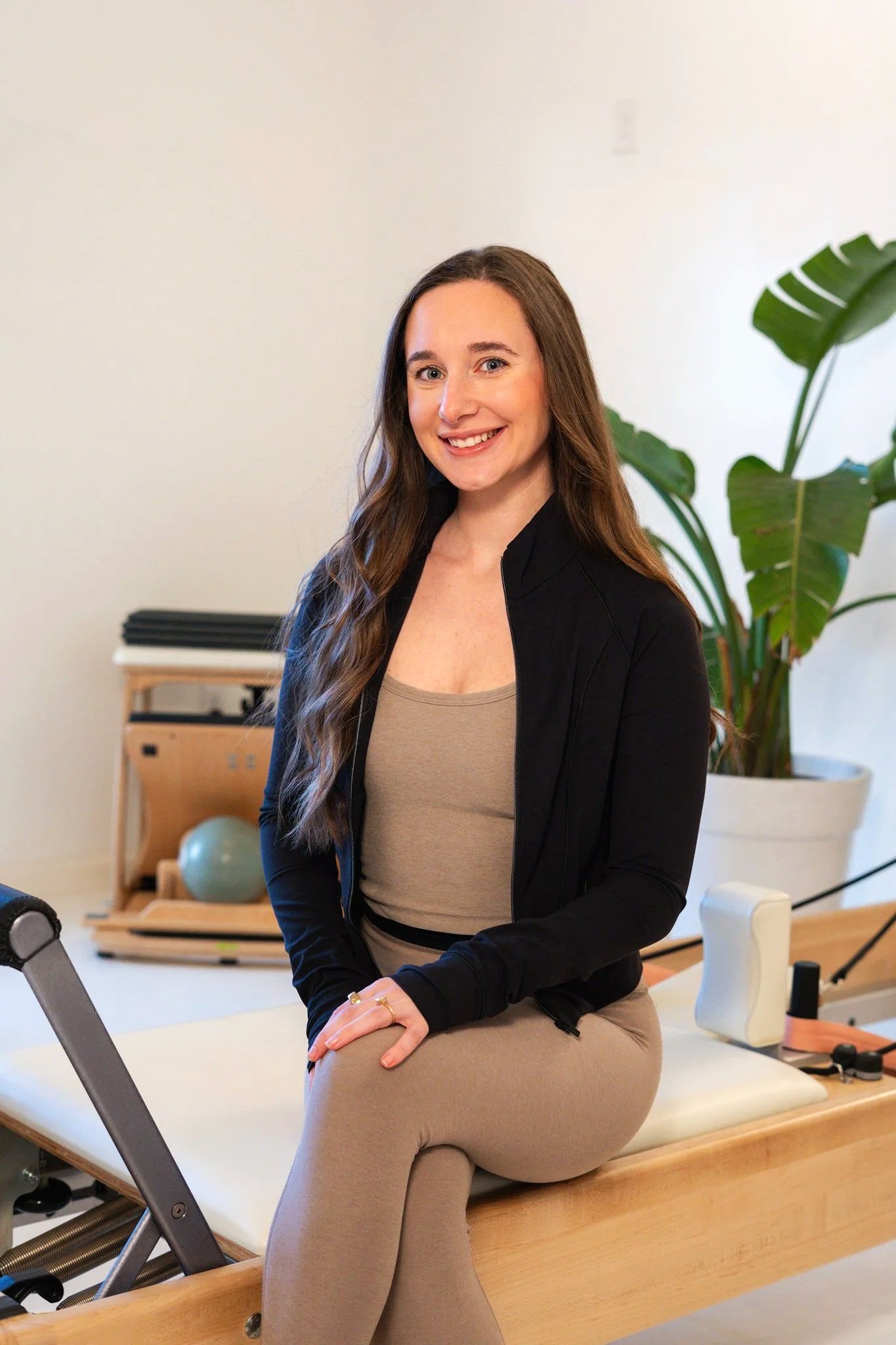 Person smiling in front of large green plants, wearing a white shirt with the text "Life Happens. Pilates Helps."