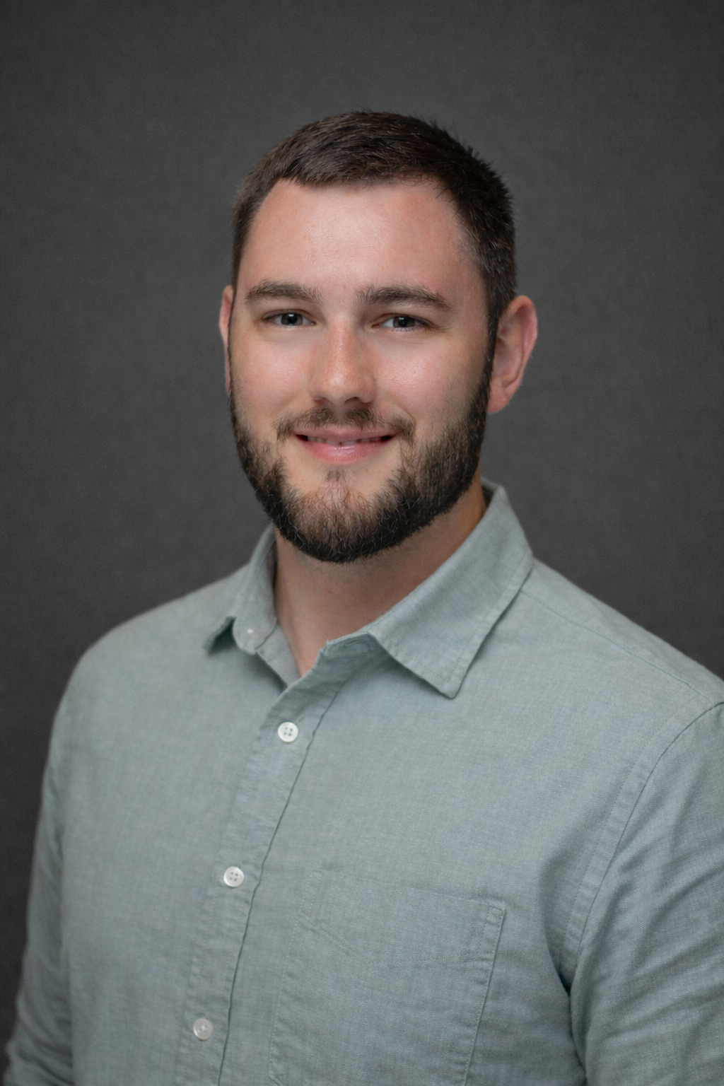 Headshot of a young man with short dark hair and a beard, wearing a light gray button-up shirt, smiling, against a dark gray background.