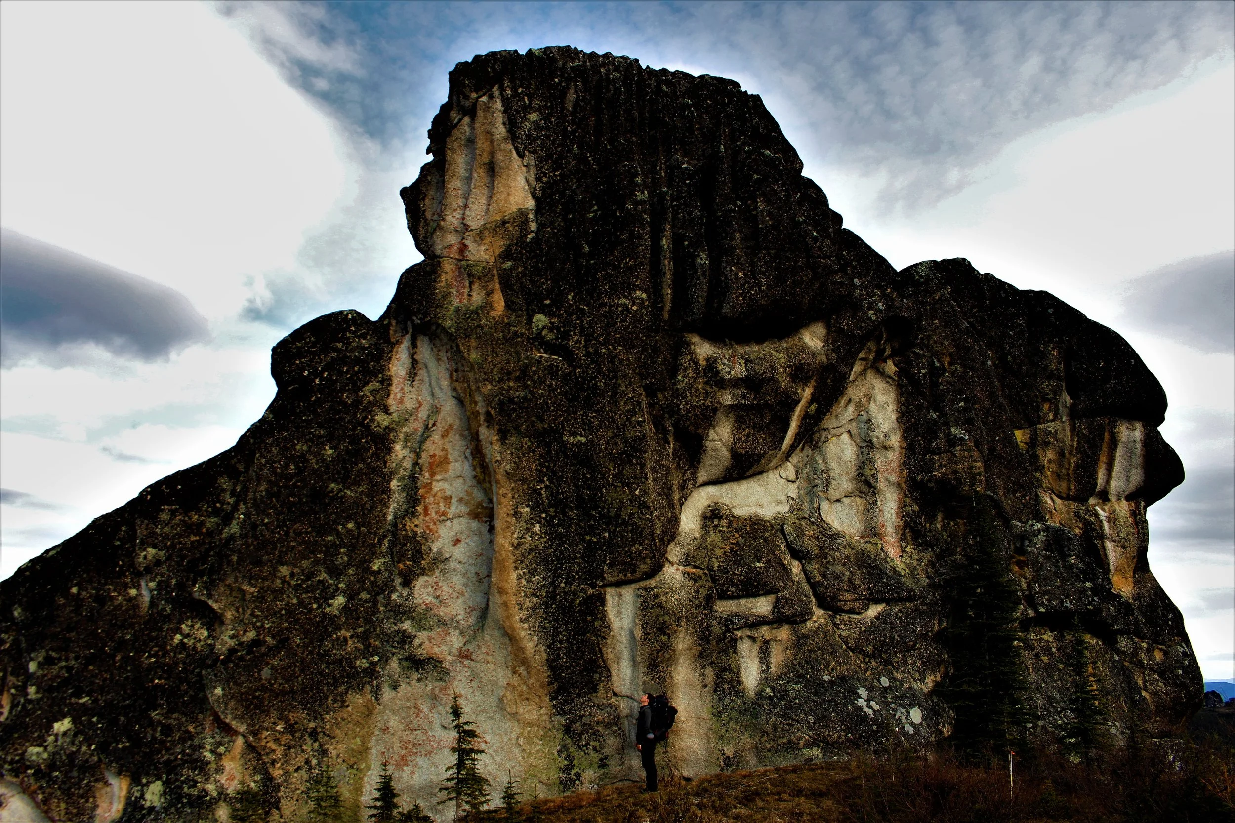 People hiking near a large granite mountain with a partly cloudy sky in the background.
