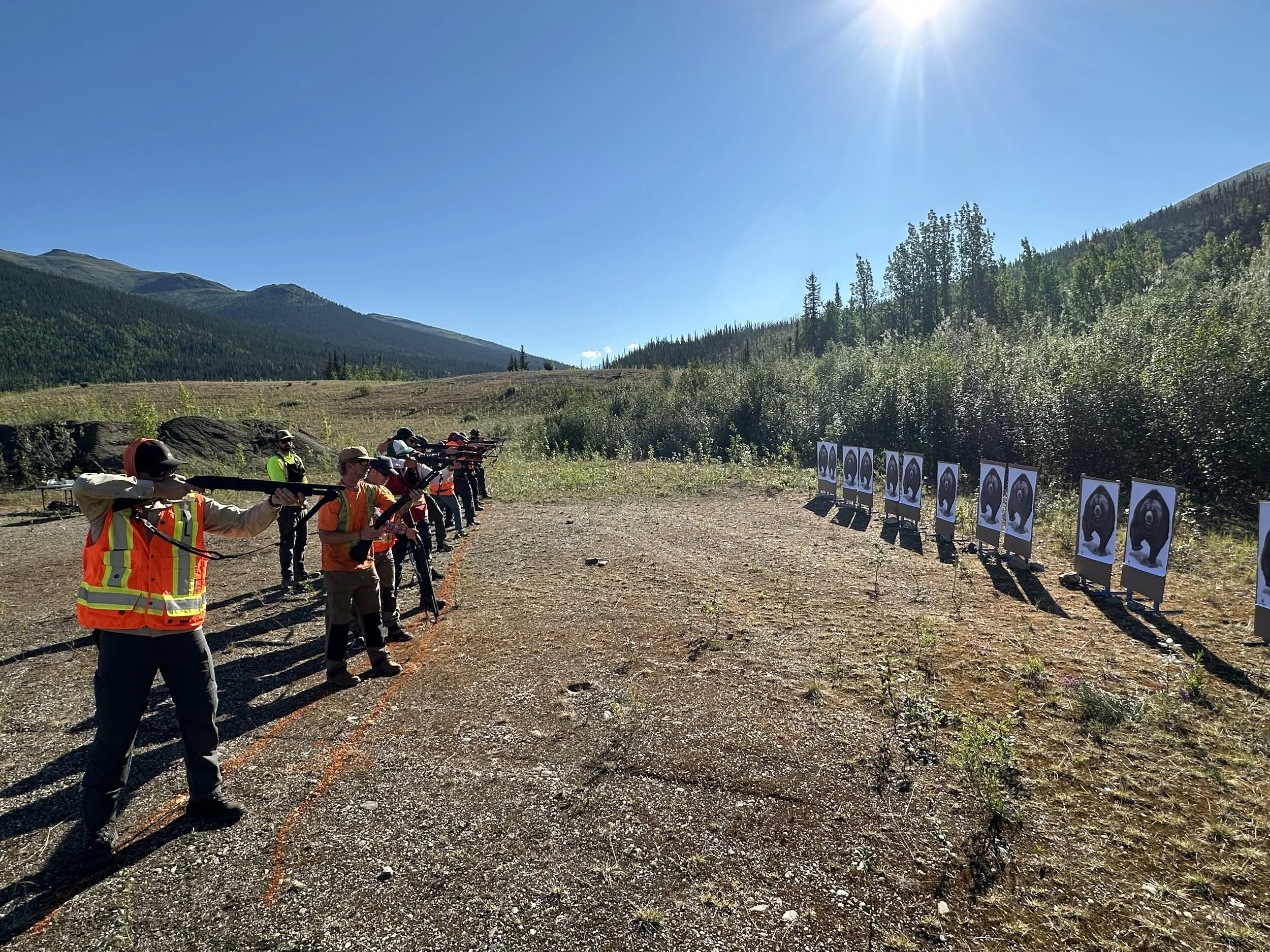 Group of people participating in a shooting activity at outdoor firing range with images of a bear on targets, situated in a mountainous forested area under bright sunlight.