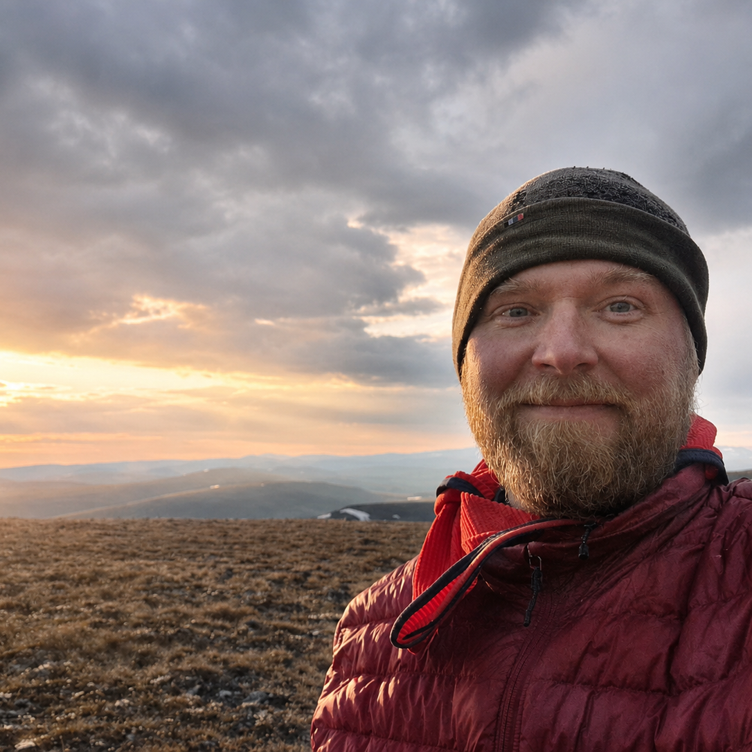A man with a beard and blue eyes taking a selfie outdoors during sunset, wearing a black beanie and a red jacket, with a landscape of rolling hills and a cloudy sky in the background.
