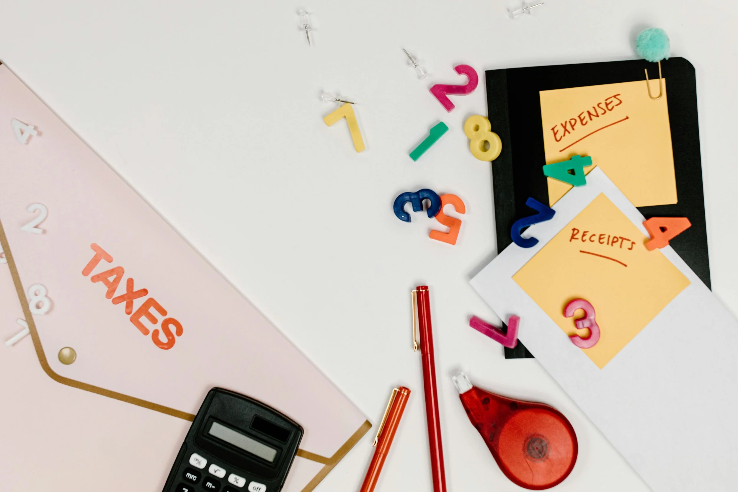 Office desk with tax organizer, calculator, pens, paper clips, and colorful plastic numbers. There are receipts and expenses notes on sticky notes and papers, indicating financial tasks.
