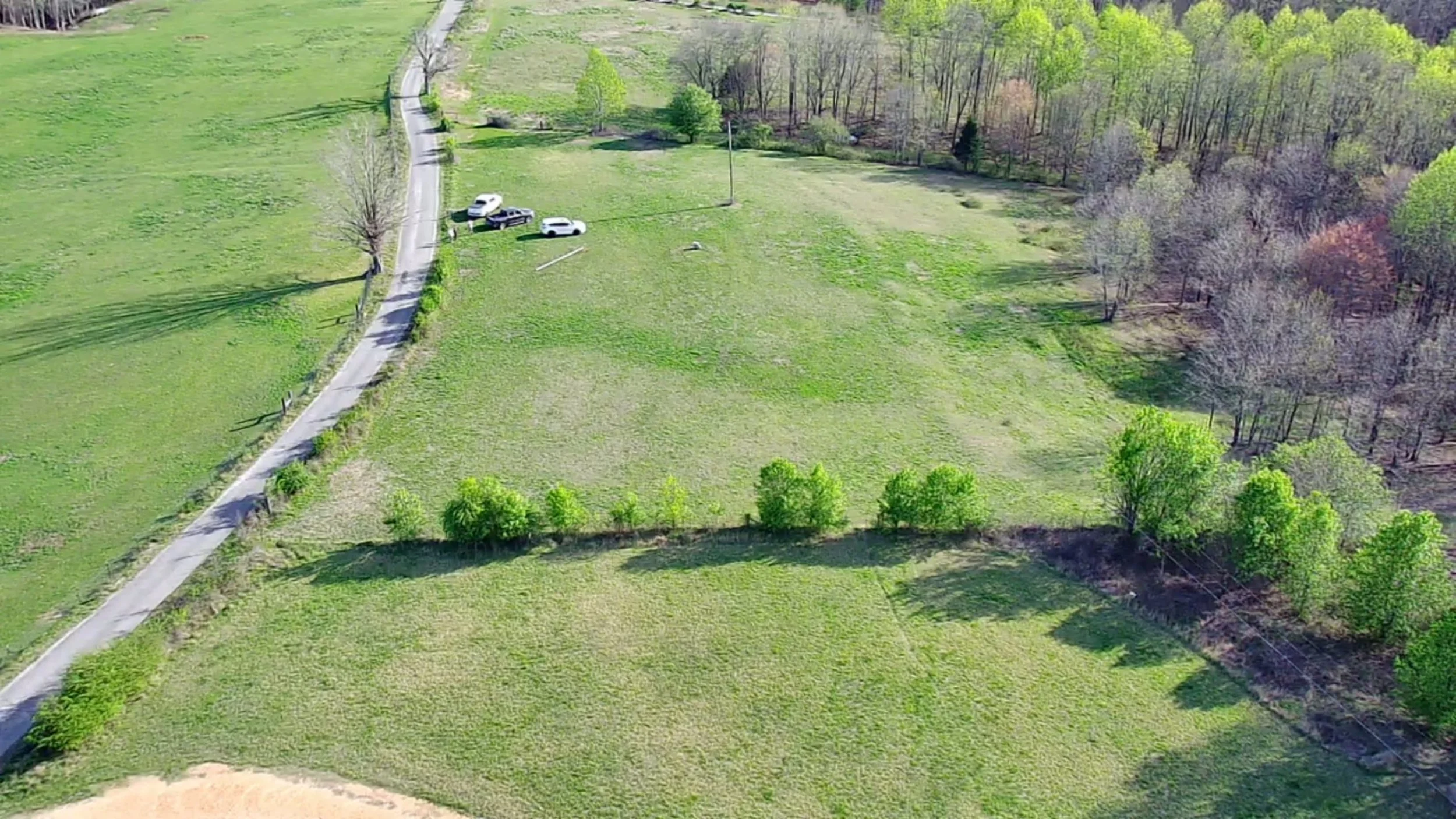 An aerial view of a grassy park with a paved path running through it, several trees scattered throughout, and a few parked cars near the path.