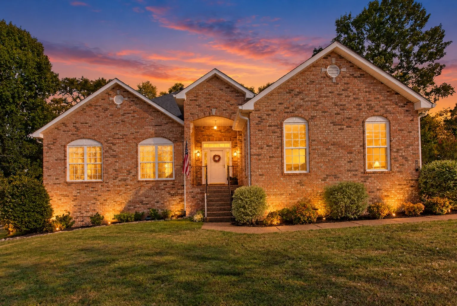 A brick house with tall windows and a front porch illuminated at sunset. There is an American flag near the steps and landscaped bushes along the front yard.