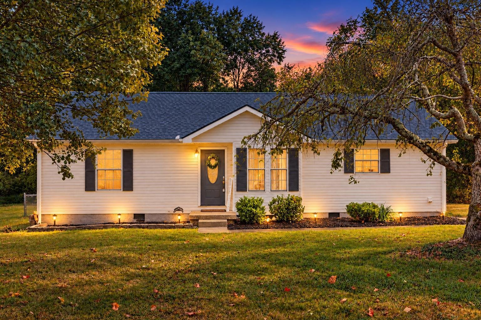 A single-story white house with black shutters and a dark door, illuminated by outdoor lights, surrounded by a green lawn and trees, during sunset.