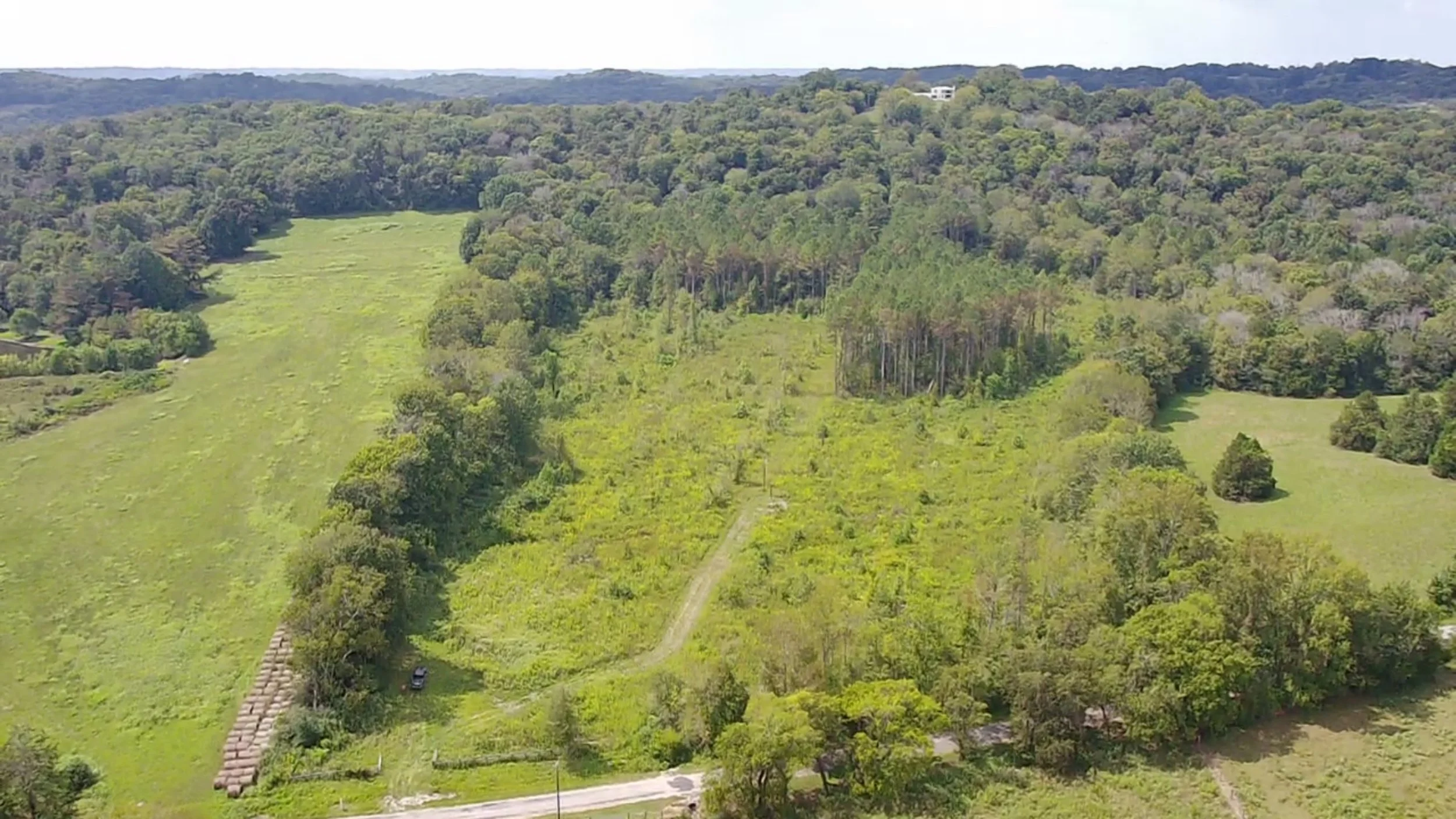 Aerial view of a lush green landscape with rolling hills, trees, open fields, and a small structure with a pathway leading to it.