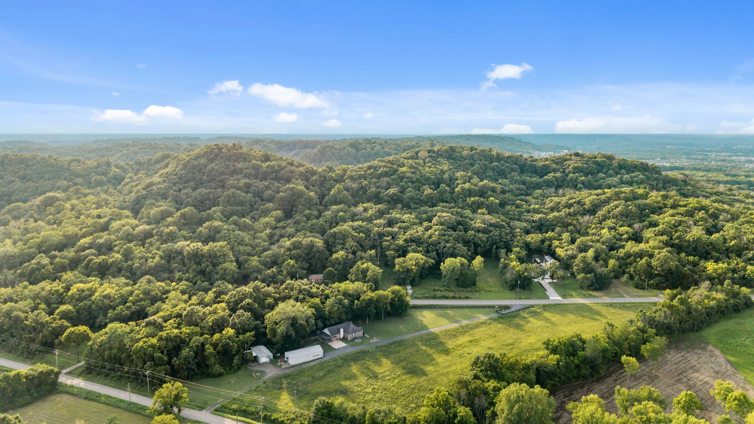 Aerial view of green forested hills with some houses, roads, and open grassy areas, under a blue sky with scattered clouds.