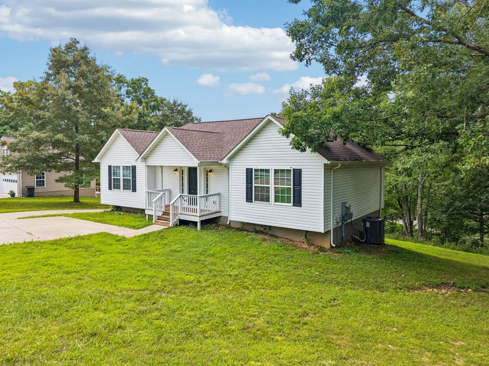 A white house with black shutters, a front porch, and a pitched roof, surrounded by green grass and large trees under a partly cloudy blue sky.
