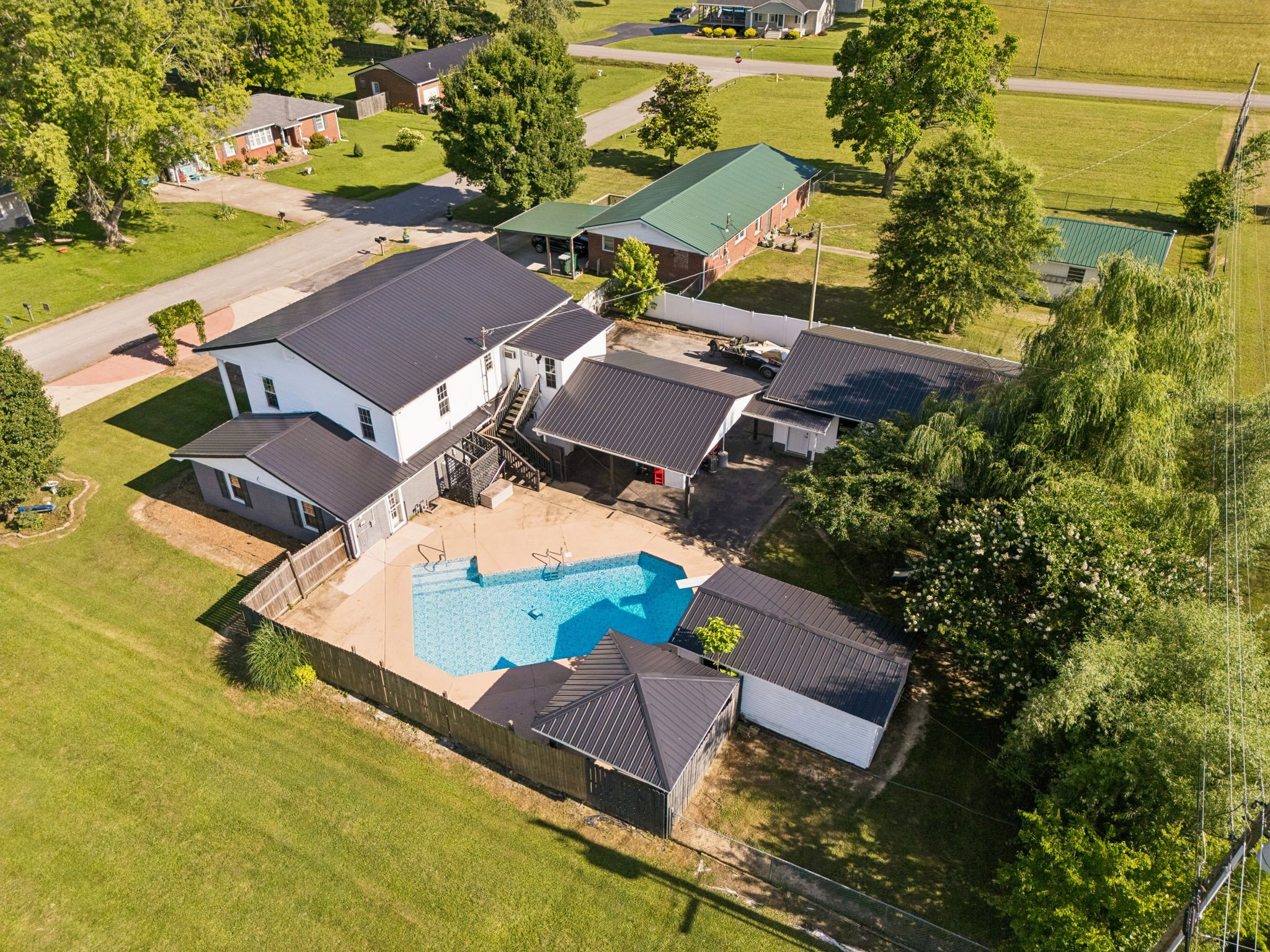 Aerial view of a suburban neighborhood featuring several houses with metal roofs, a swimming pool, green lawns, and trees.