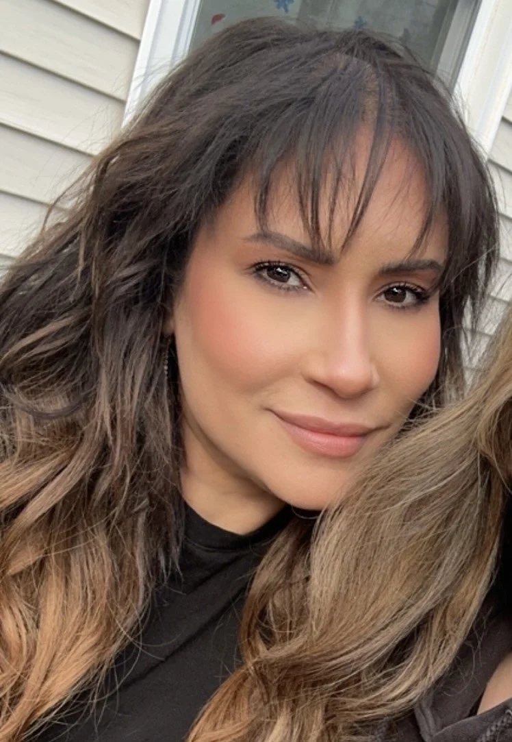 Close-up of a smiling woman with long wavy hair and bangs, wearing a black top, outdoors.