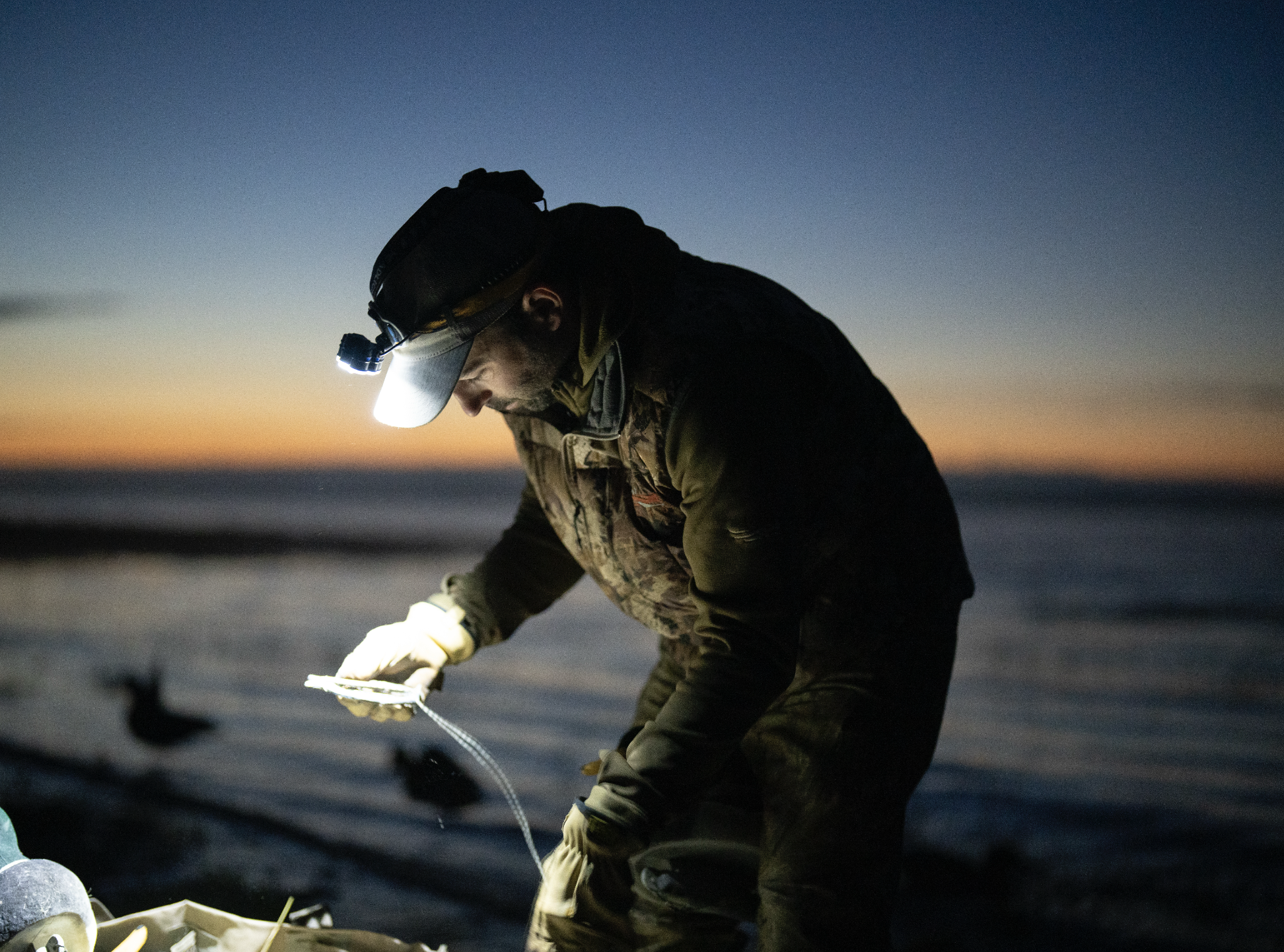Personne portant une lampe frontale examinant un objet près de l'eau au crépuscule. Person wearing headlamp examining item near water at dusk