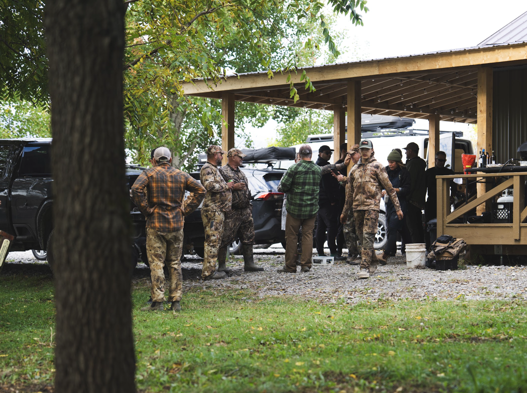 Un groupe de personnes portant des vêtements de camouflage et de plein air rassemblé sous un abri en bois près de véhicules stationnés dans un cadre extérieur verdoyant.