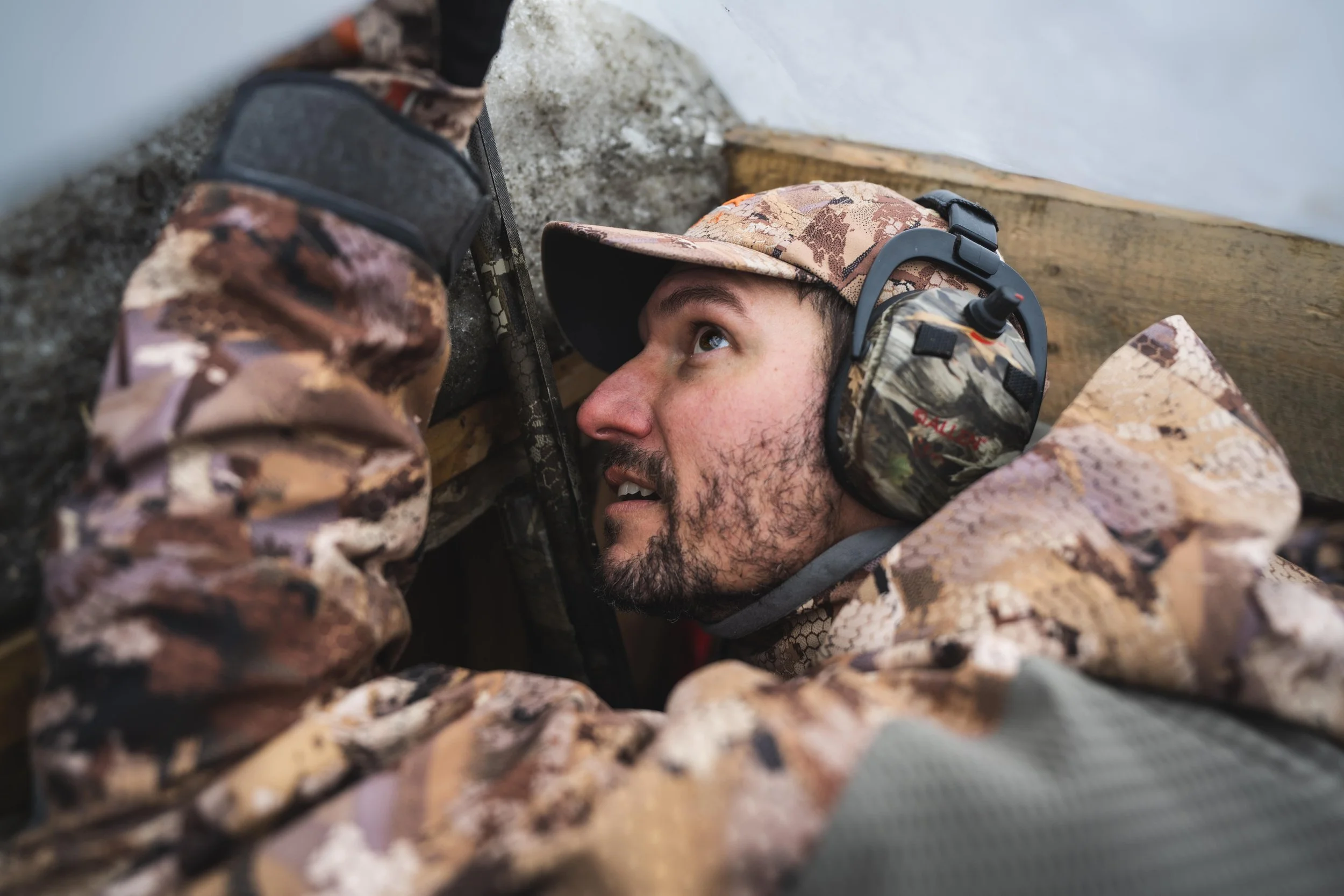 Un homme en tenue de camouflage, portant une protection auditive, regarde vers le haut, positionné dans une zone abritée en plein air. Man in camouflage gear wearing ear protection looks up, positioned in a sheltered area outdoors.