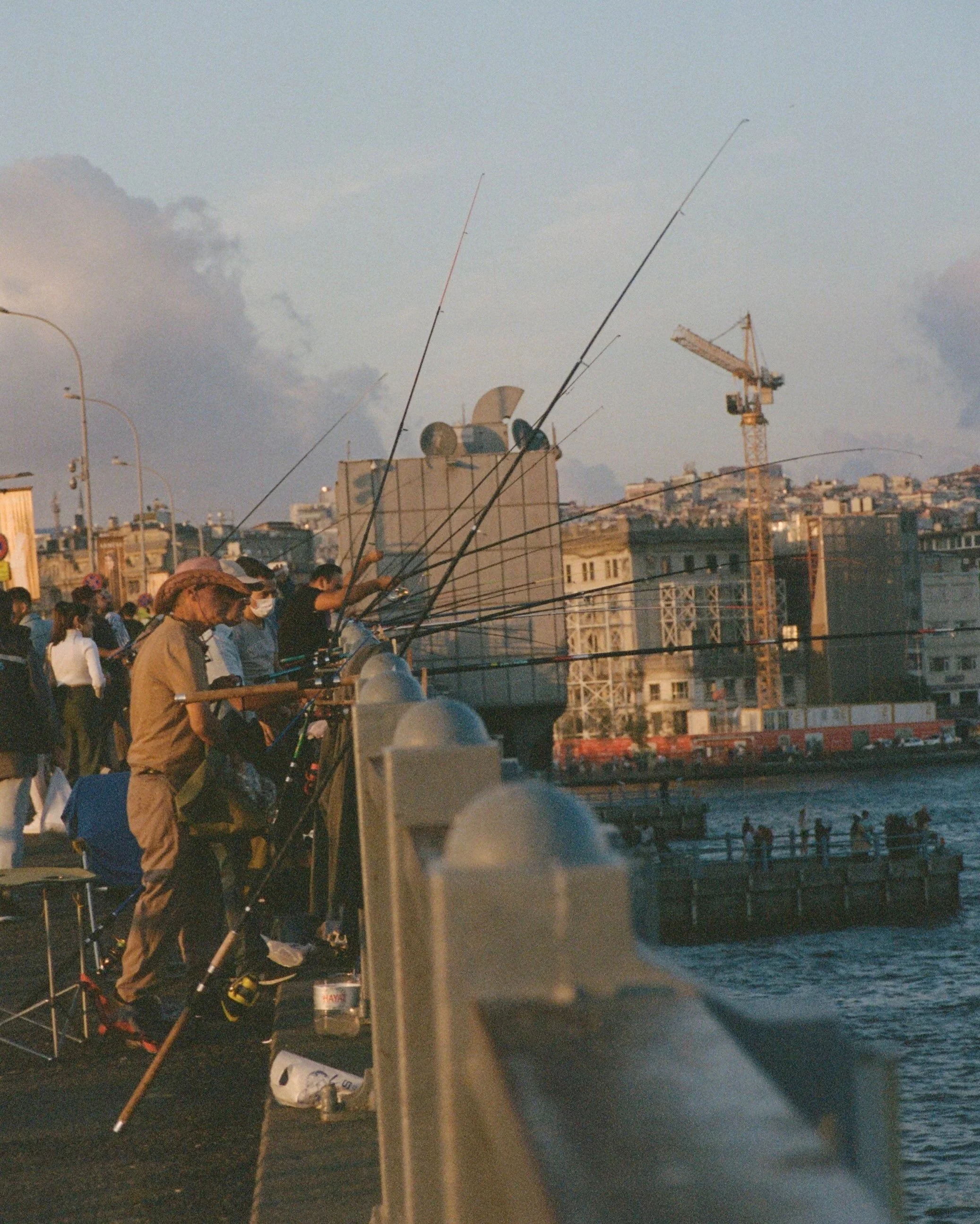 People fishing off a city pier at sunset with city buildings and a construction crane in the background.
