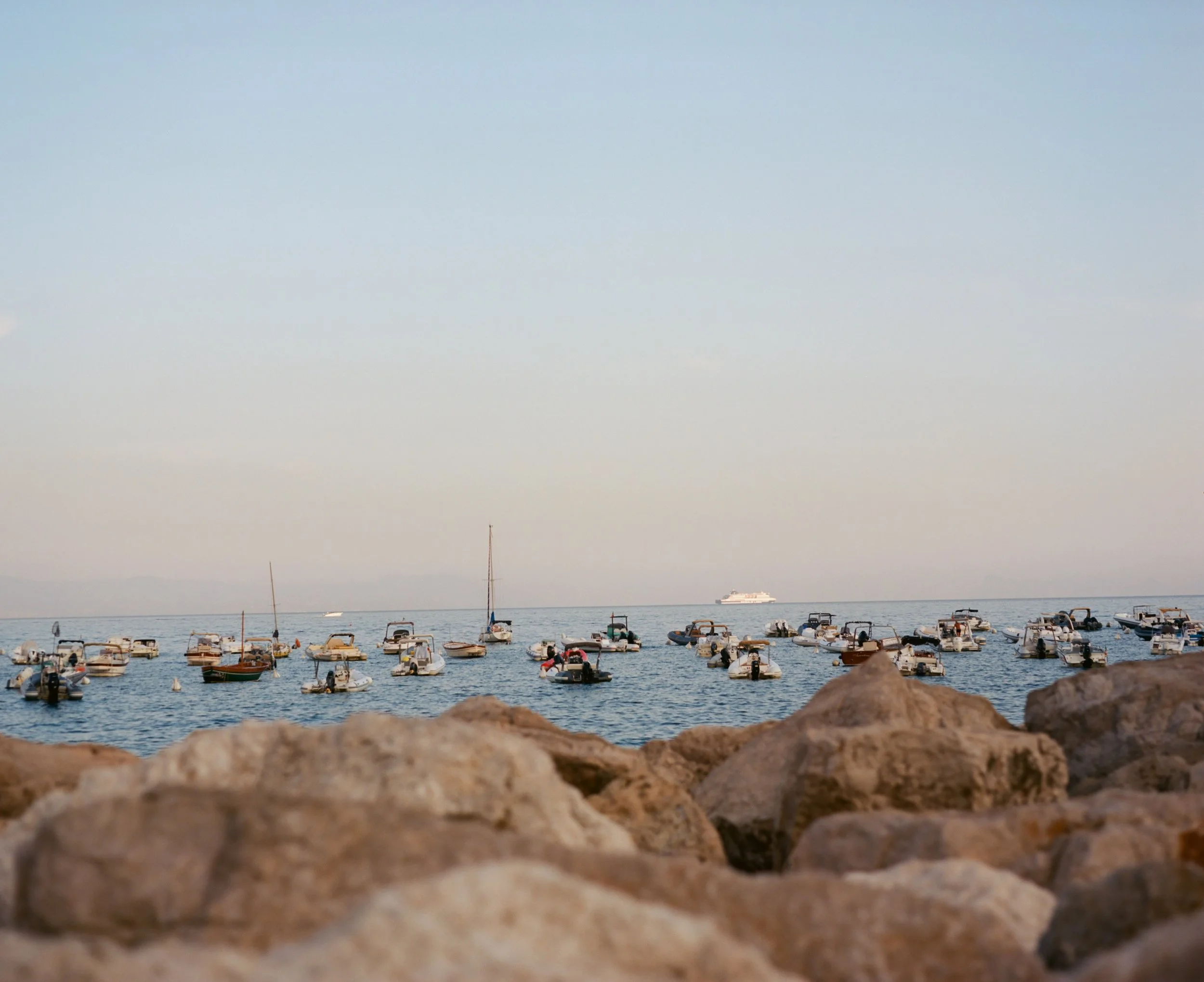 Multiple small boats anchored near the rocky shoreline on calm water, with a large ship in the background under a clear sky.