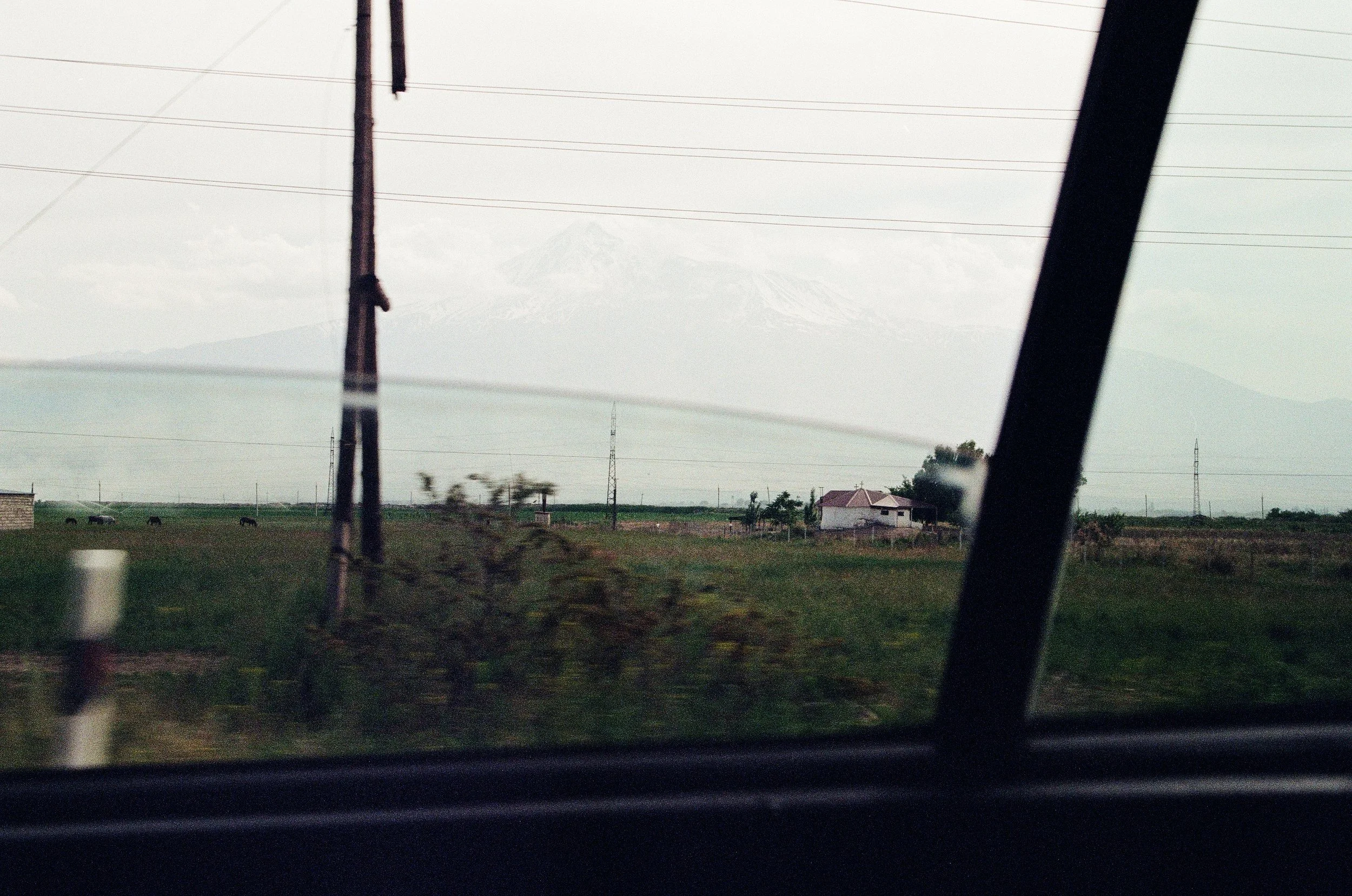 View of a distant snow-capped mountain through a car window, with power lines and a small house in the foreground.