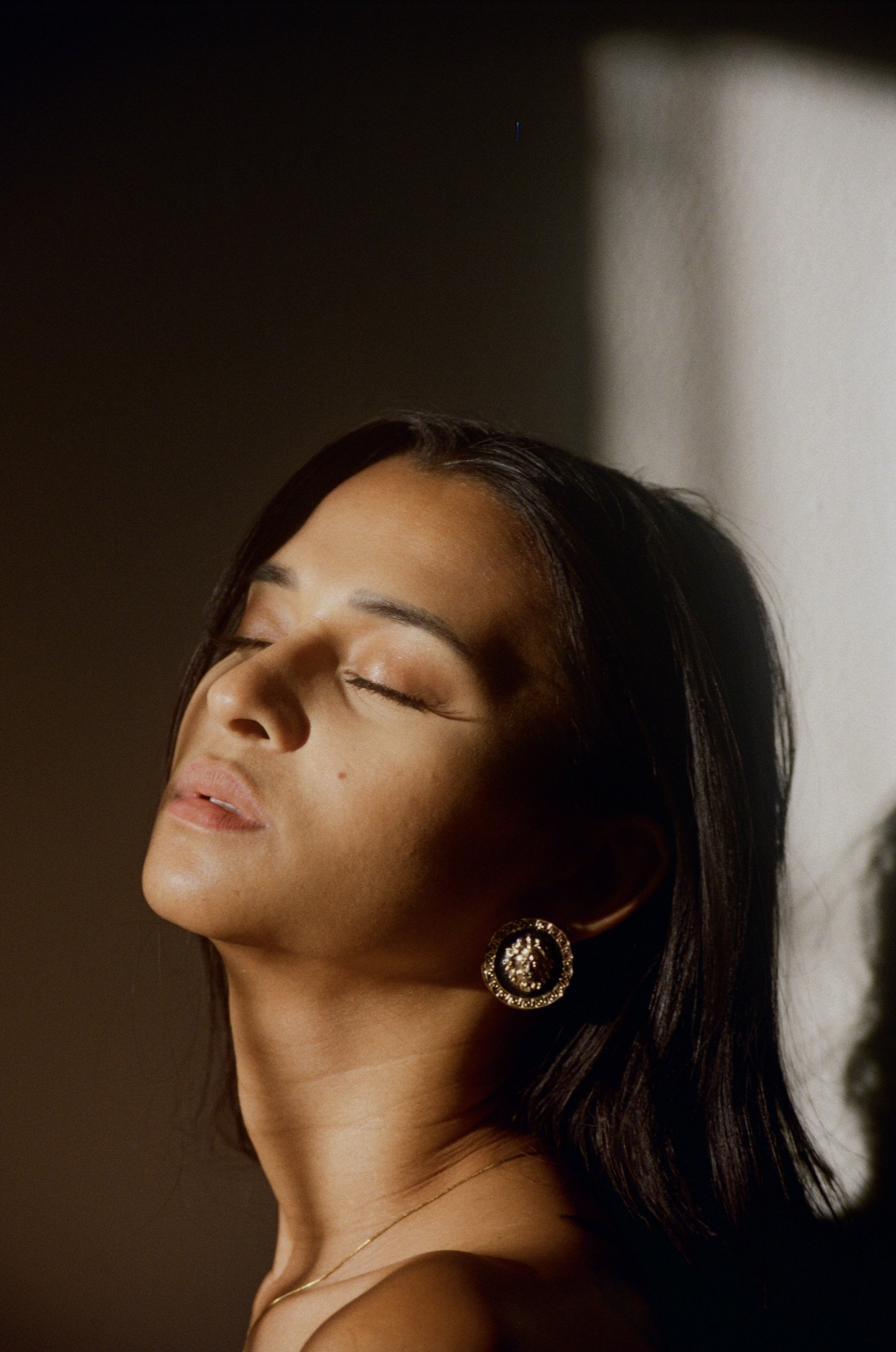 Close-up of a woman with dark hair and gold earrings, her eyes closed, basking in sunlight with a serene expression.
