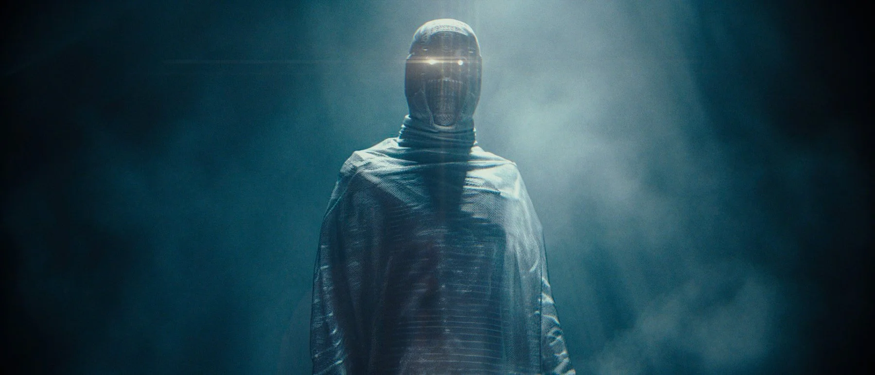 Person wearing a metallic or futuristic helmet and cloak, standing in a misty, dark environment with a backlight creating a glowing effect around the head.