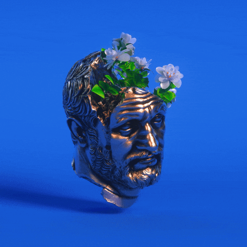 A metallic bust of a bearded man with a crown of white flowers and green leaves on his head, against a blue background.