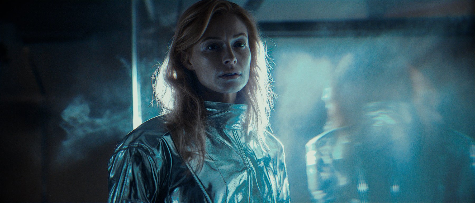 A woman with long red hair, wearing a shiny metallic silver jacket, stands in a dimly lit, smoky environment with a serious expression.