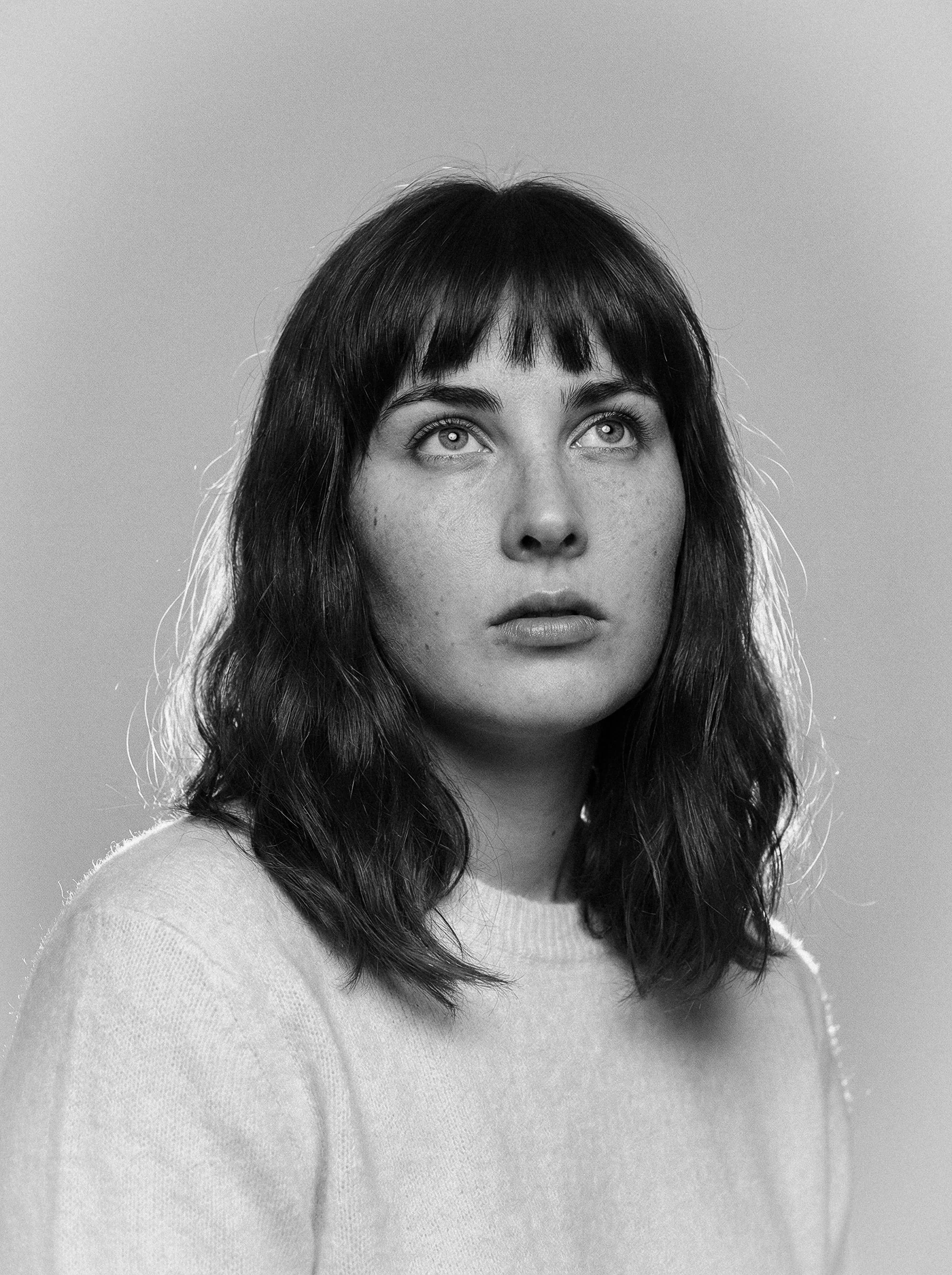 Black and white portrait of a young woman with shoulder-length wavy hair and bangs, wearing a light-colored sweater, against a plain background.