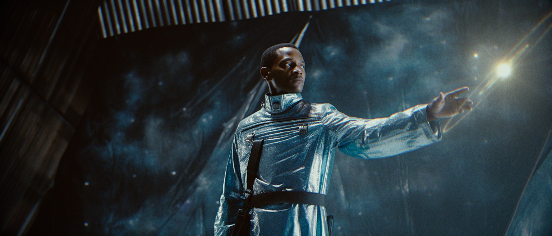 A person in a shiny silver futuristic outfit reaching out their hand towards a bright light in a space-like environment.