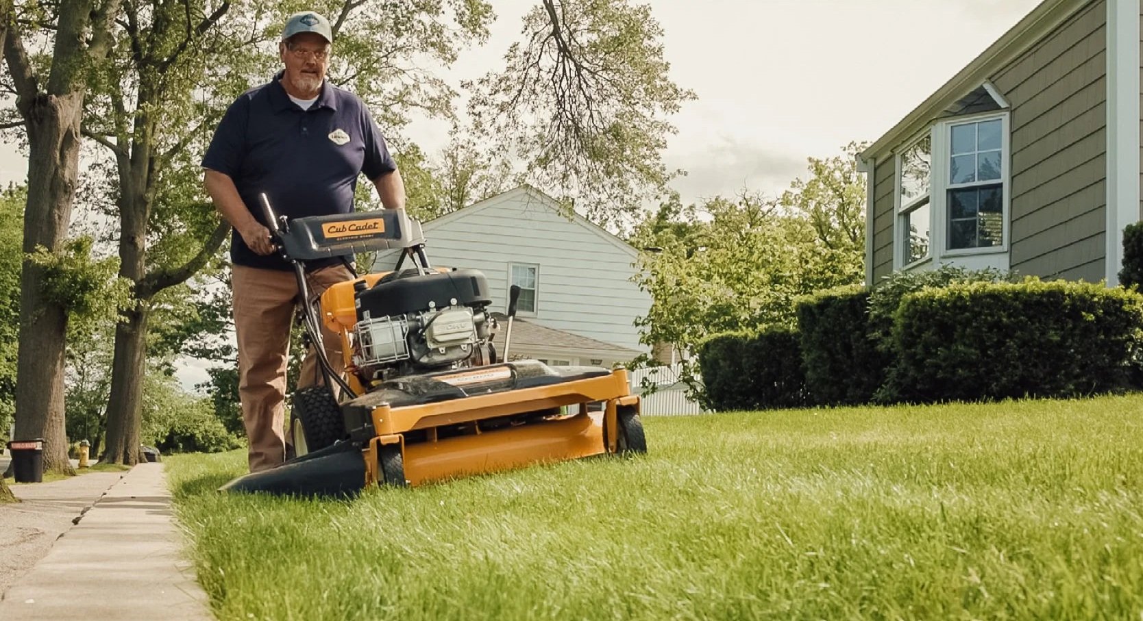 Man mowing lawn with a yellow and black mower in front of a house with green bushes and trees.